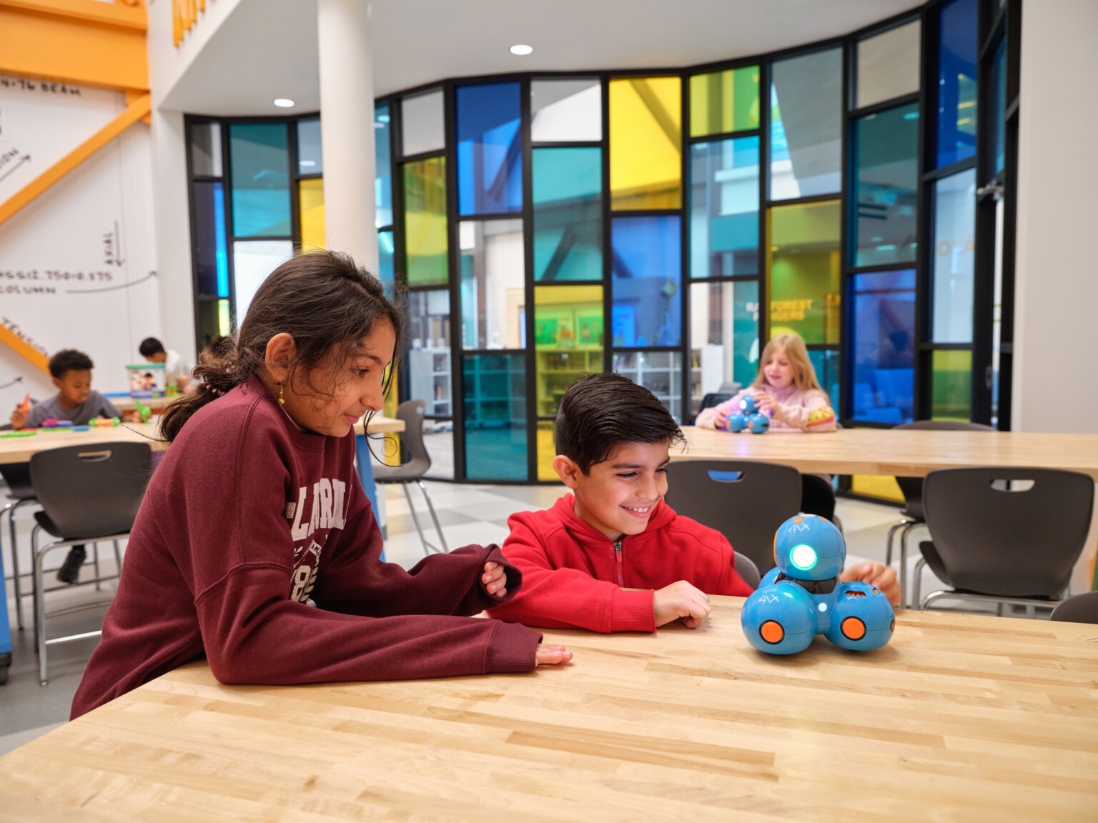 Students at Crestview Elementary School engaging with a digital toy at desks in a classroom; colorful stained window in the background; white walls with orange accents perpendicular to the colorful wall