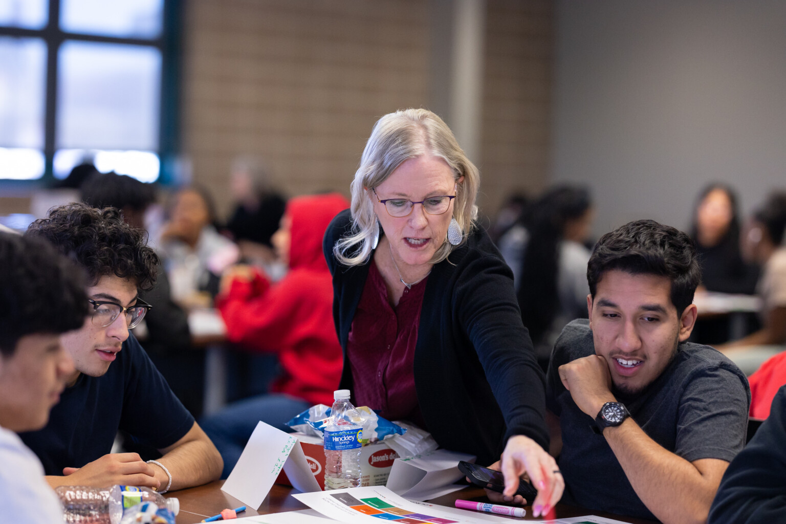 woman helping students and professionals at a table; large hall with many tables of people collaborating