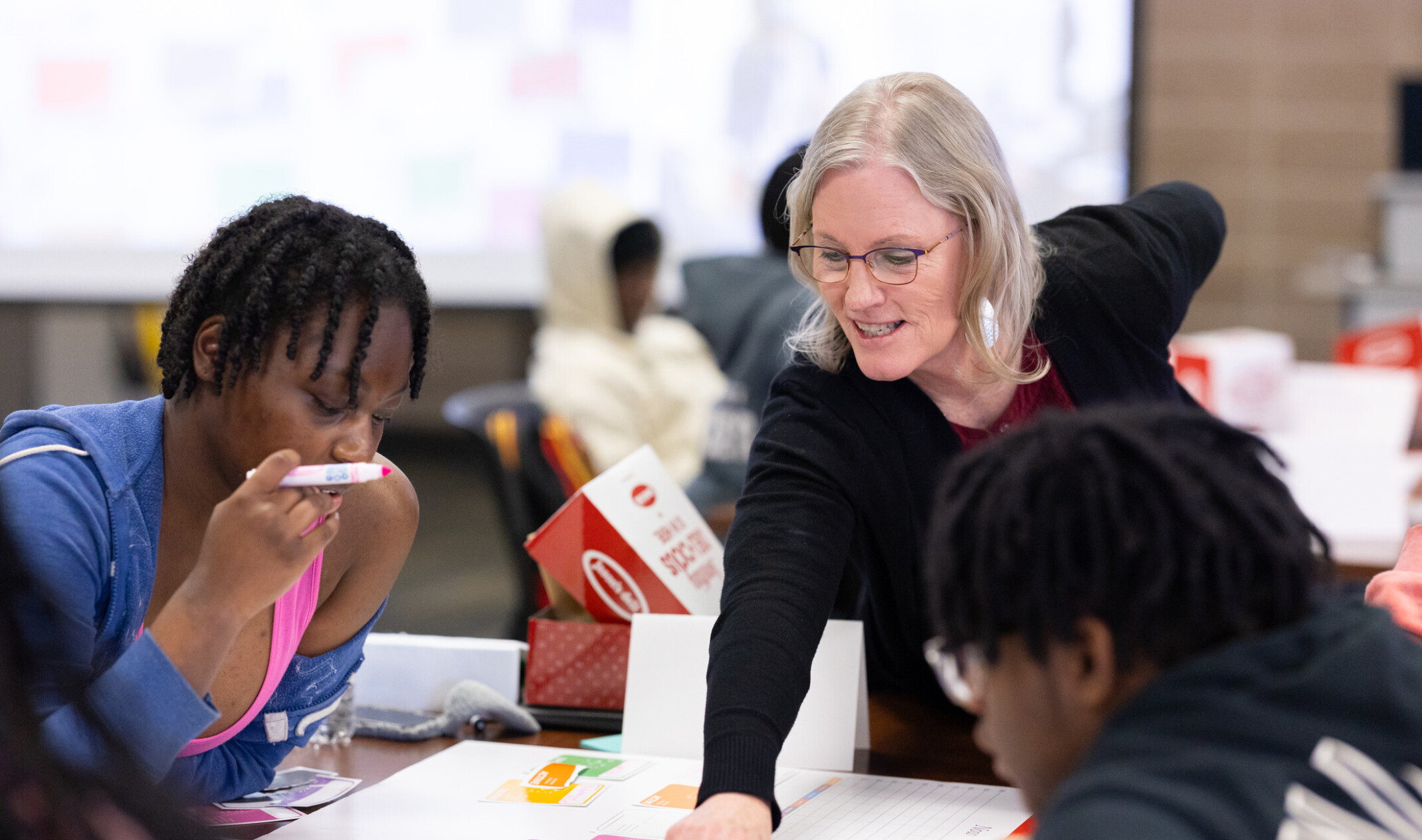 woman helping students and professionals at a table; large hall with many tables of people collaborating