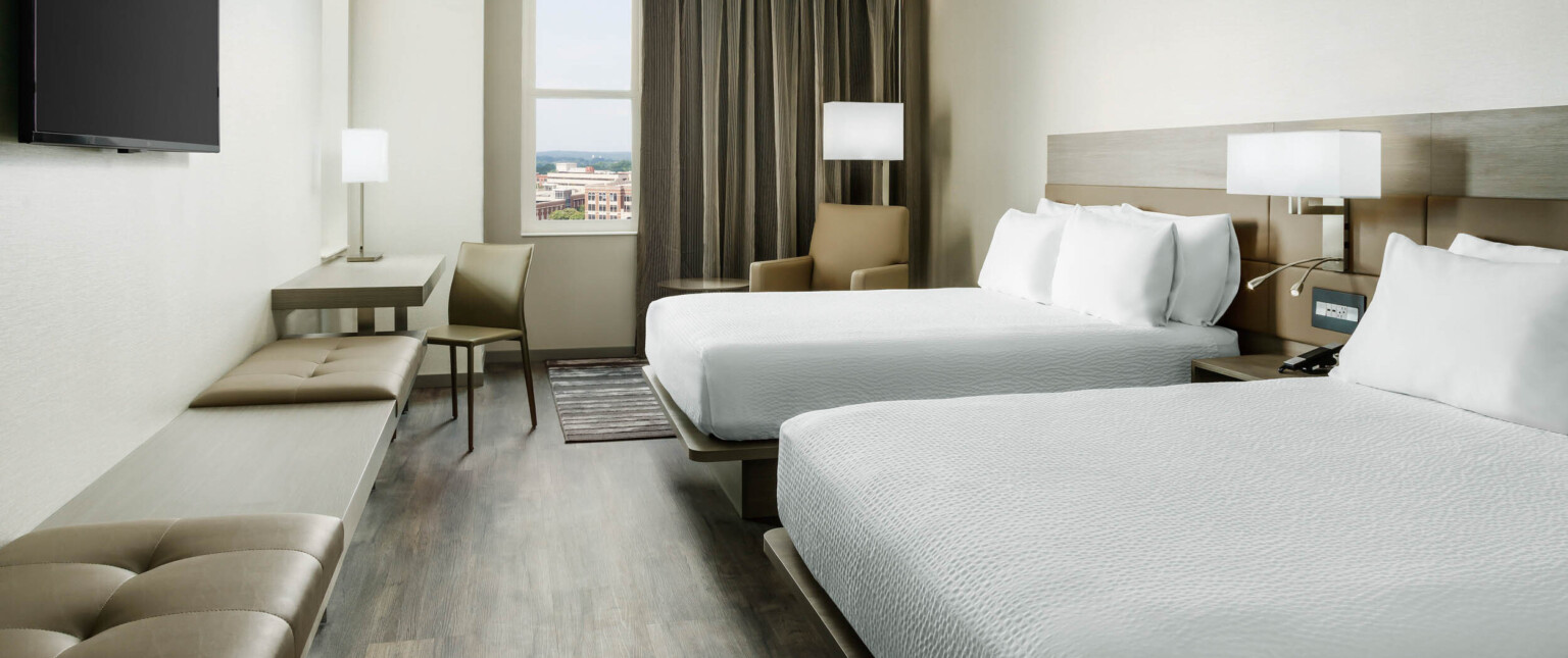 Guest room with two white beds divided by an end table and light fixture. Arm chair by window, all face bench along left wall following Marriott brand architecture