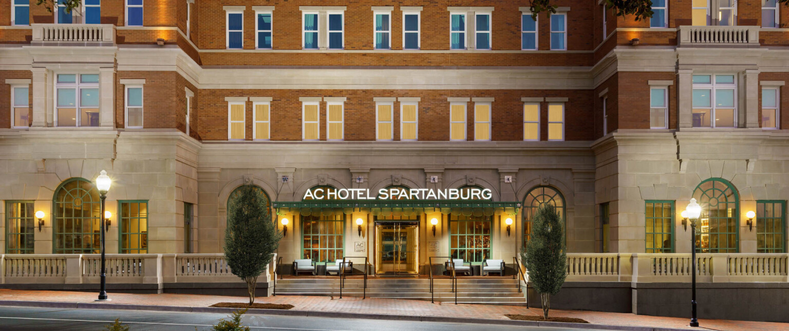 Exterior of hotel entry with sign illuminated on green awning. Green arches windows on carved front stone wall