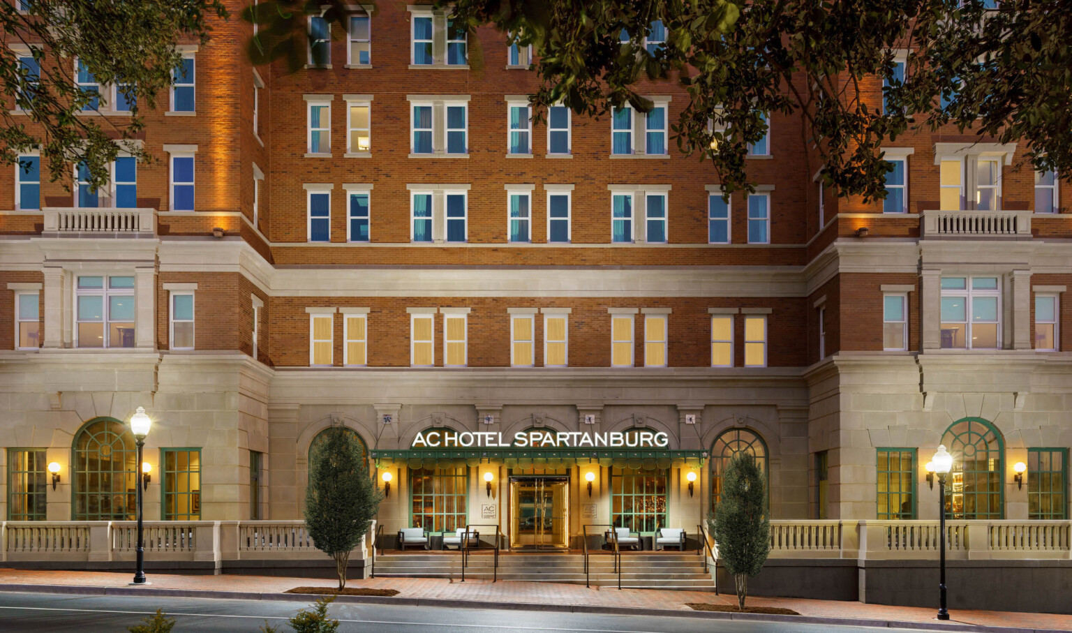 Exterior of hotel entry with sign illuminated on green awning. Green arches windows on carved front stone wall