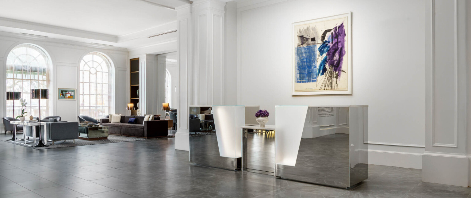 Metallic reception desk with illuminated detail in white AC Hotel Spartanburg lobby with large arch window, seating area left