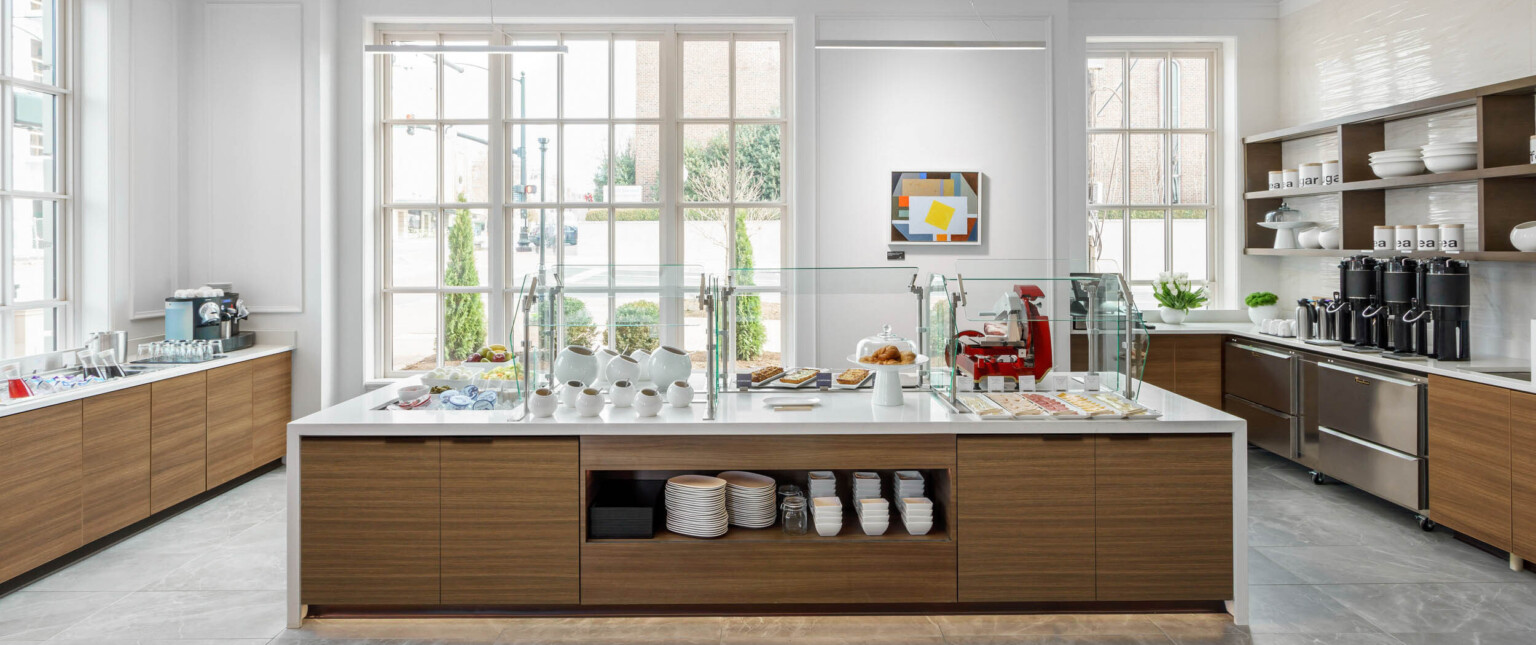 White counter with wood storage underneath set for a breakfast buffet in a white room with large windows on ground level