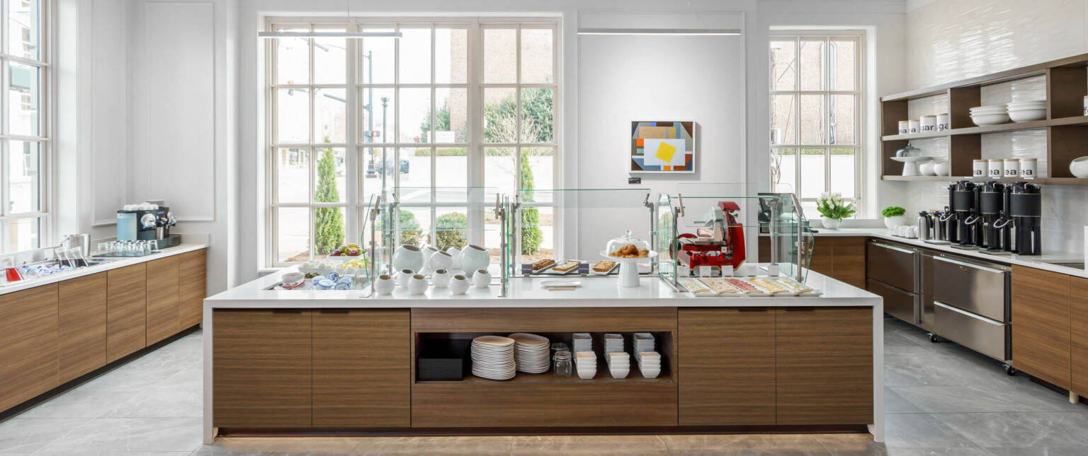 White counter with wood storage underneath set for a breakfast buffet in a white room with large windows on ground level following Marriott brand architecture