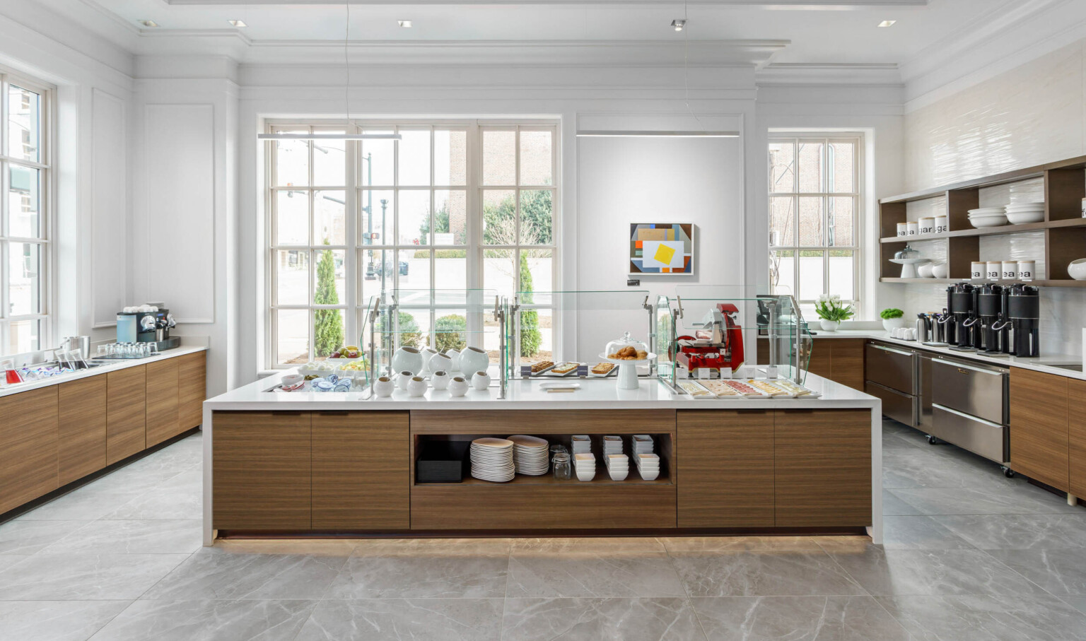 White counter with wood storage underneath set for a breakfast buffet in a white room with large windows on ground level