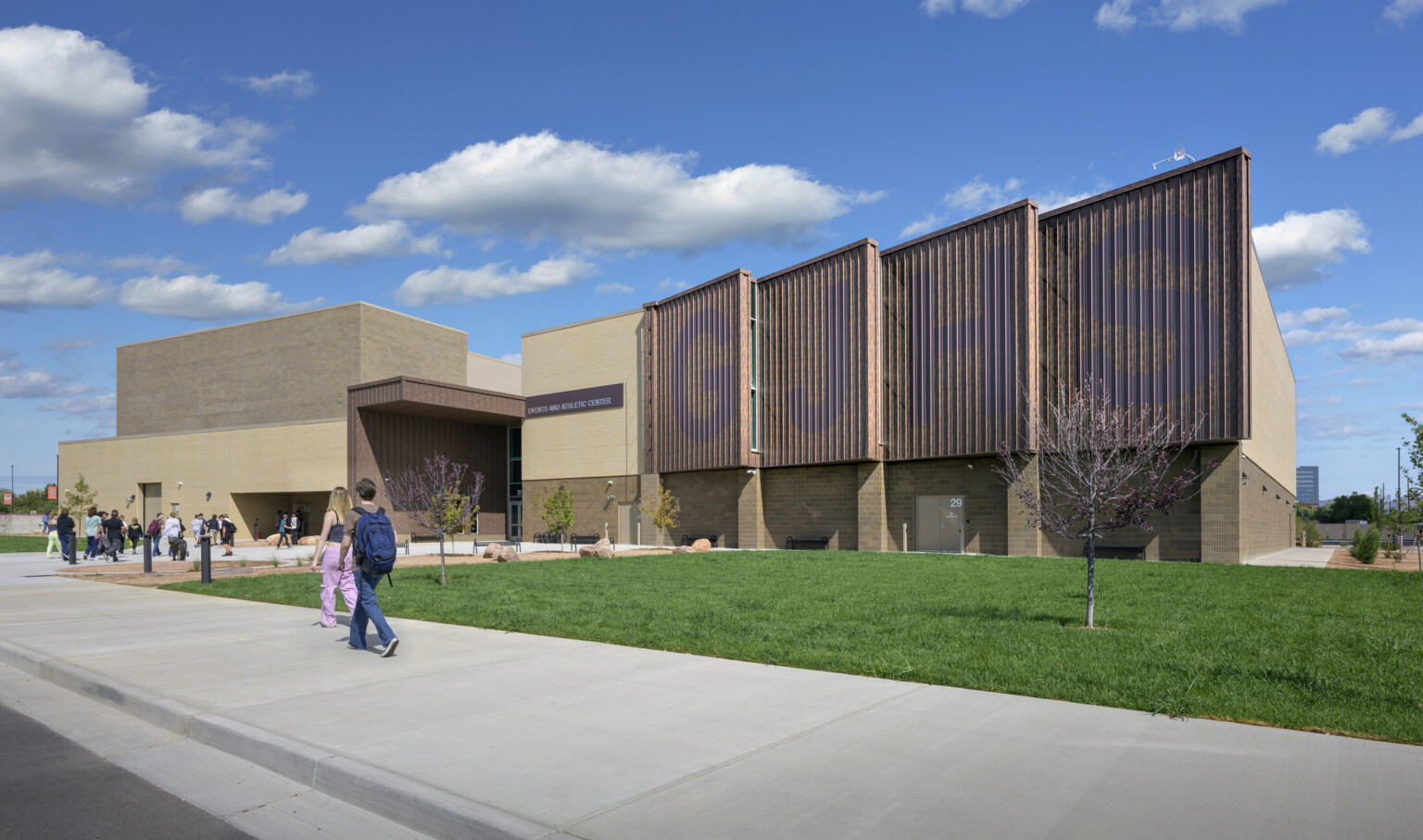 Front facing image of Grand Junction High School with students walking in; slated front of the building has large GJHS painted in blue on facade; tan and brown building stands in front of sparsely cloudy blue sky