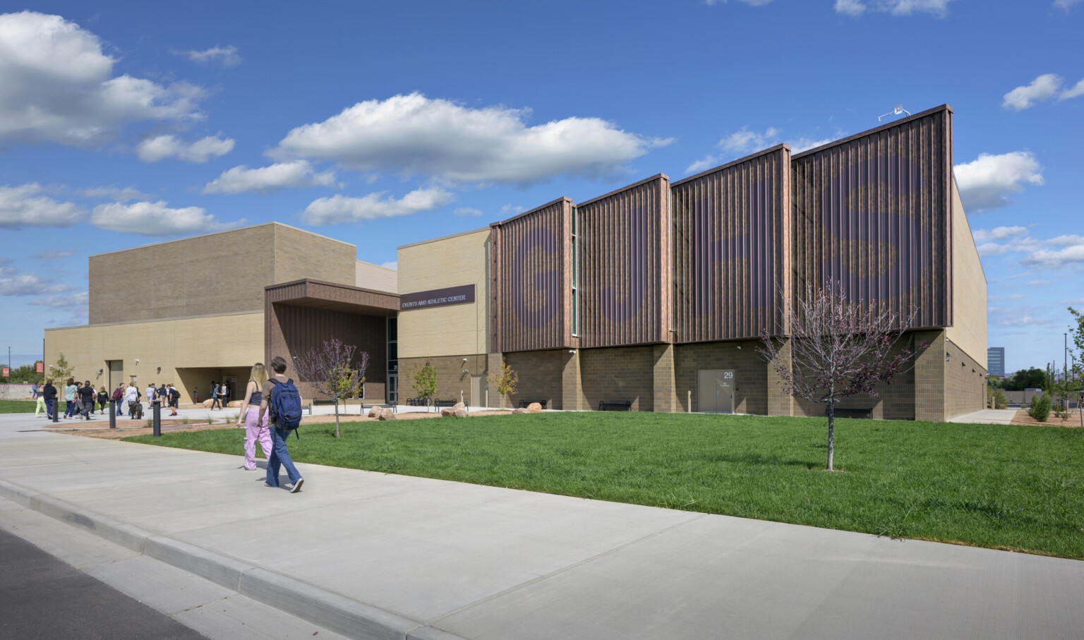 Front facing image of Grand Junction High School with students walking in; slated front of the building has large GJHS painted in blue on facade; tan and brown building stands in front of sparsely cloudy blue sky