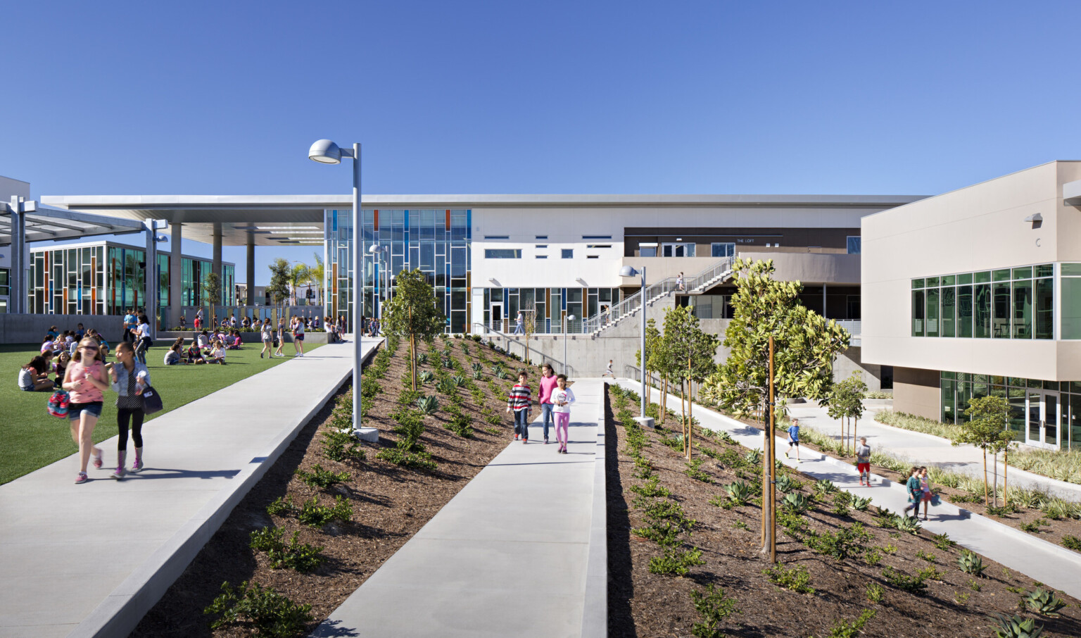 children walking on diagonal concrete paths between landscaping in front of Design 39 campus, a multistory building with glass façade; non traditional school campus with indoor and outdoor spaces