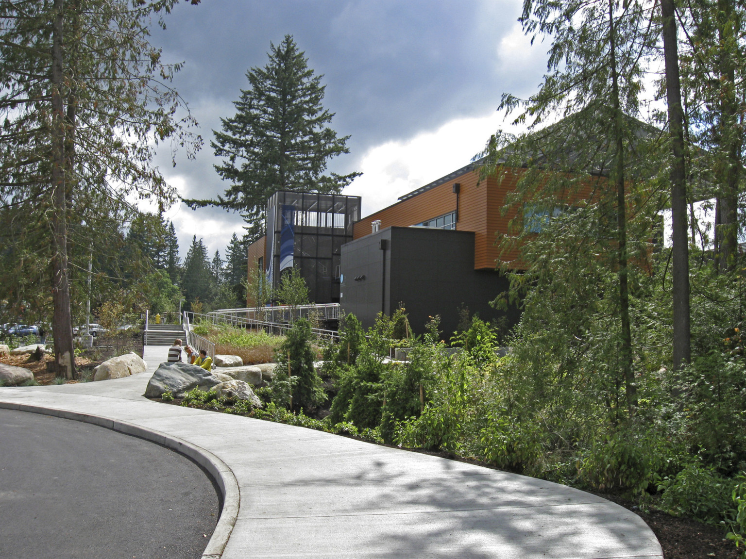 Tall narrow trees and shrubs along rounded road with sidewalk leading to orange and grey building with angled roof panel