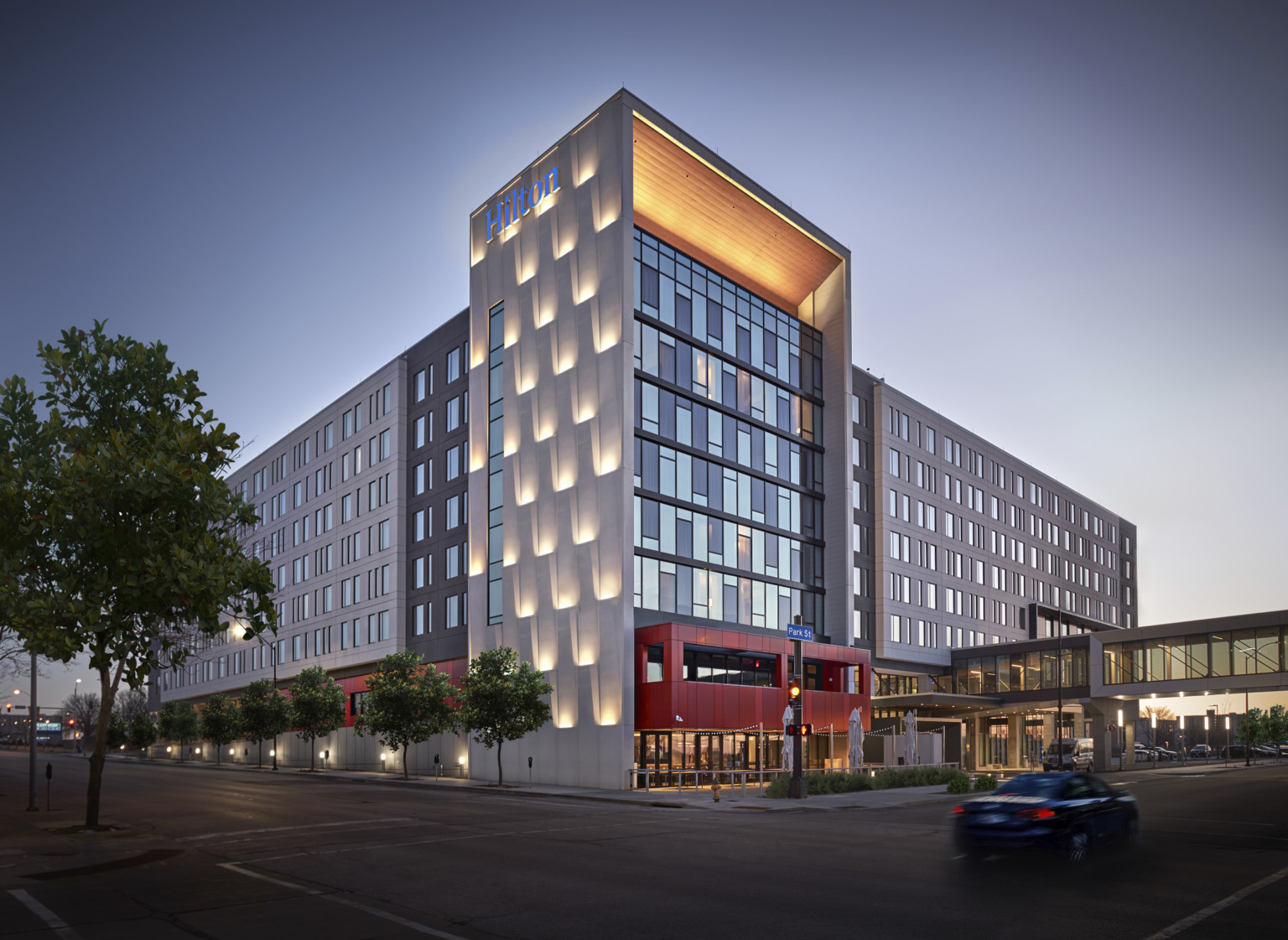 exterior corner view in the evening of multistory hotel with lit up roof arch, warm red entryway, and scaled facade with uplighting at hilton des moines