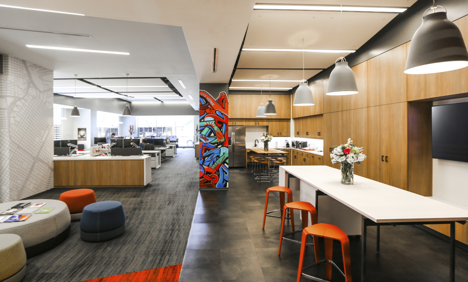 Office space with wood panel kitchen to right with counter height tables. Mural on center column, desks and seating to left Office space with wood panel kitchen to right with counter height tables. Mural on center column, desks and seating to left
