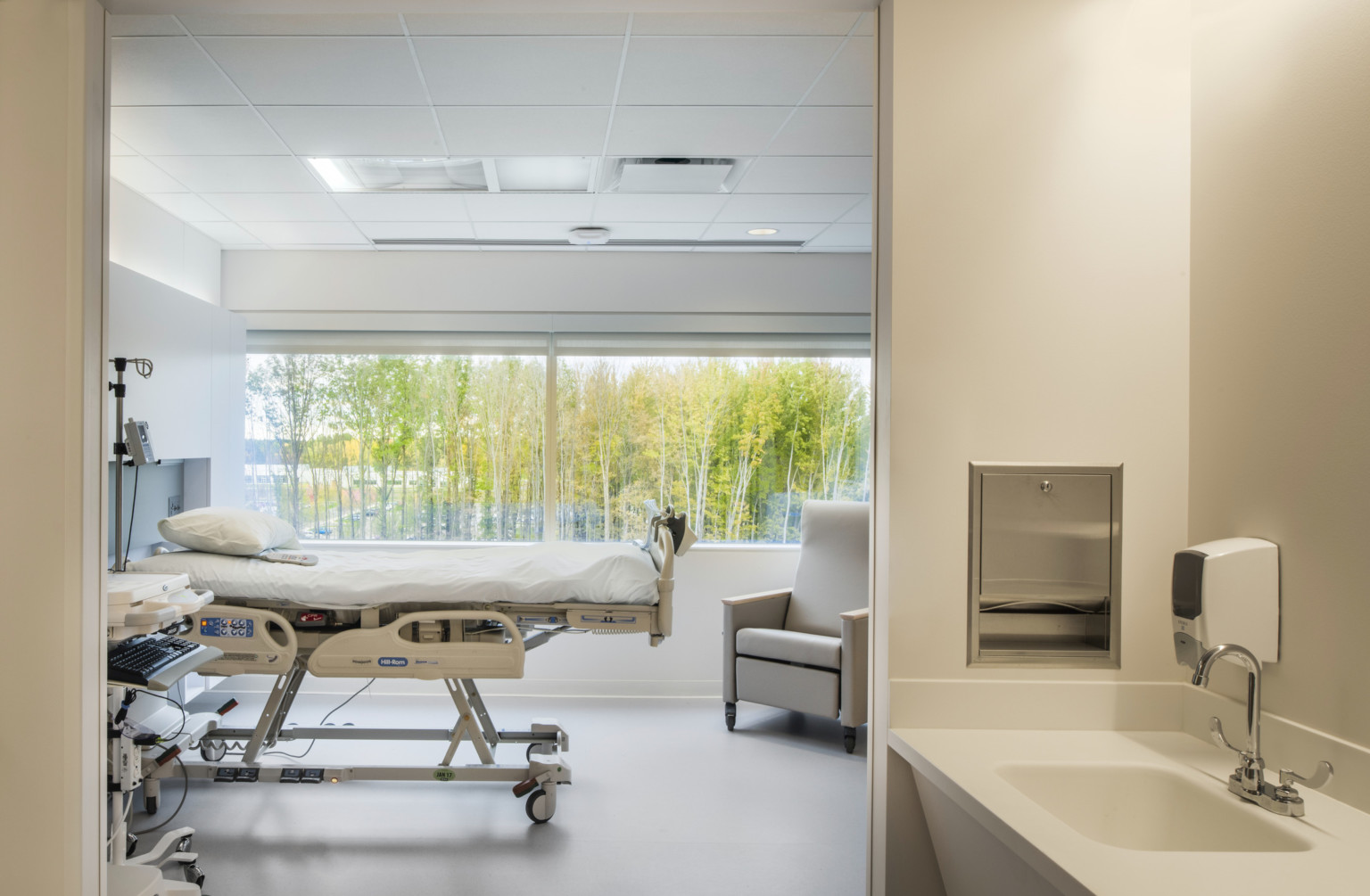 Hospital bed reclined in front of large window with views of trees. An arm chair faces the bed at the foot