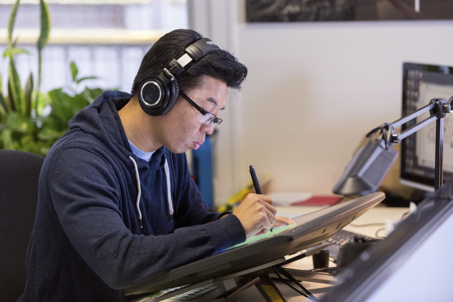 Man sketching on touchscreen with headphones on at desk in white office. Plants in window behind