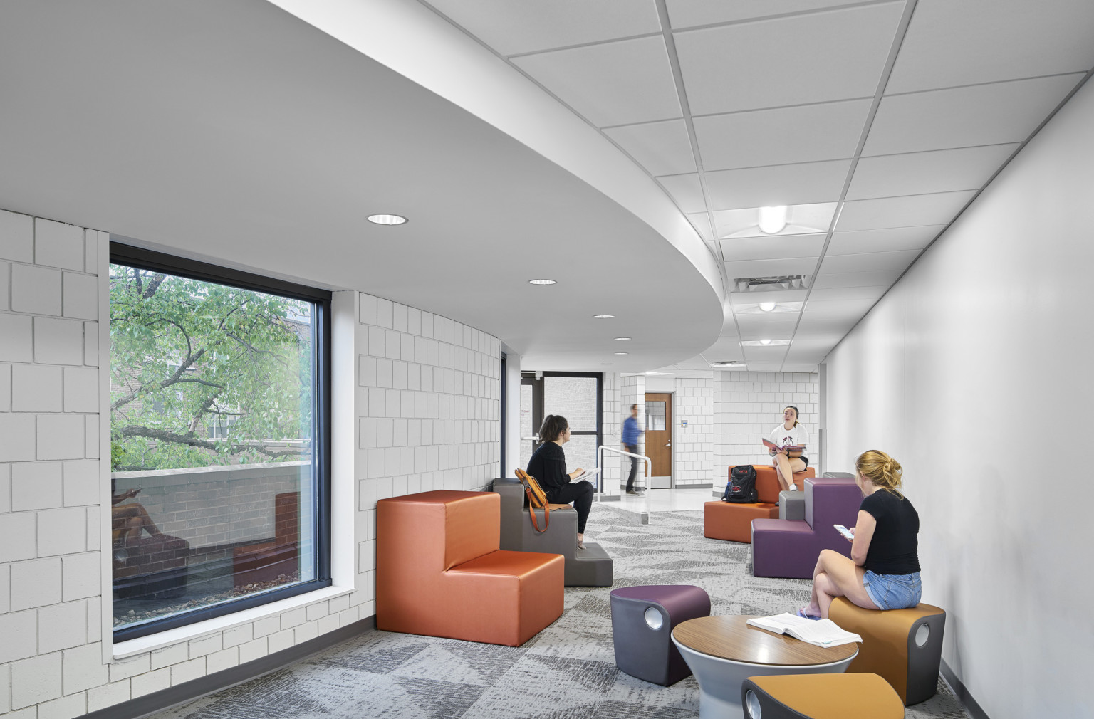 Renovated curved white tiled hallway for student interaction with colorful moveable seating. Curved drop ceiling detail, left