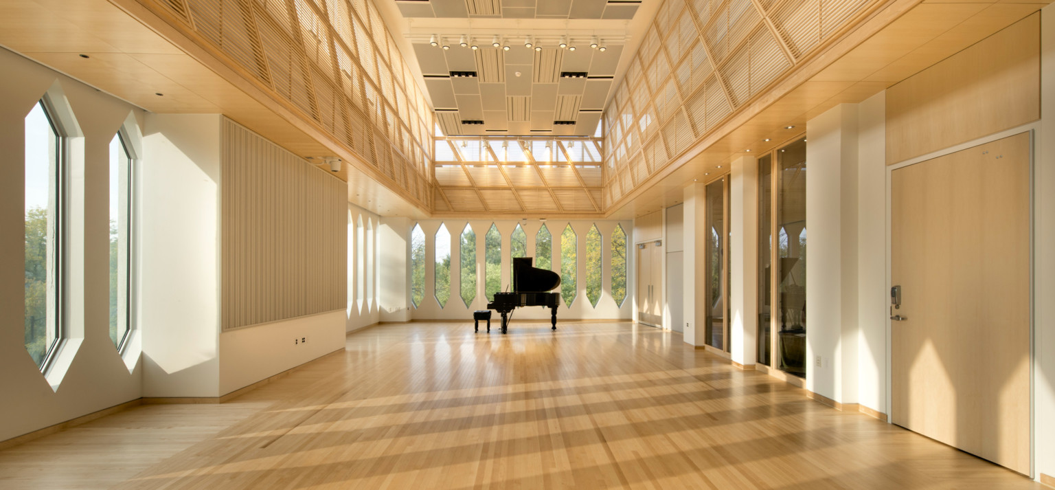 Piano in the center of a corner room with white walls and thin rectangular windows with pointed tops and bottoms, wood floor