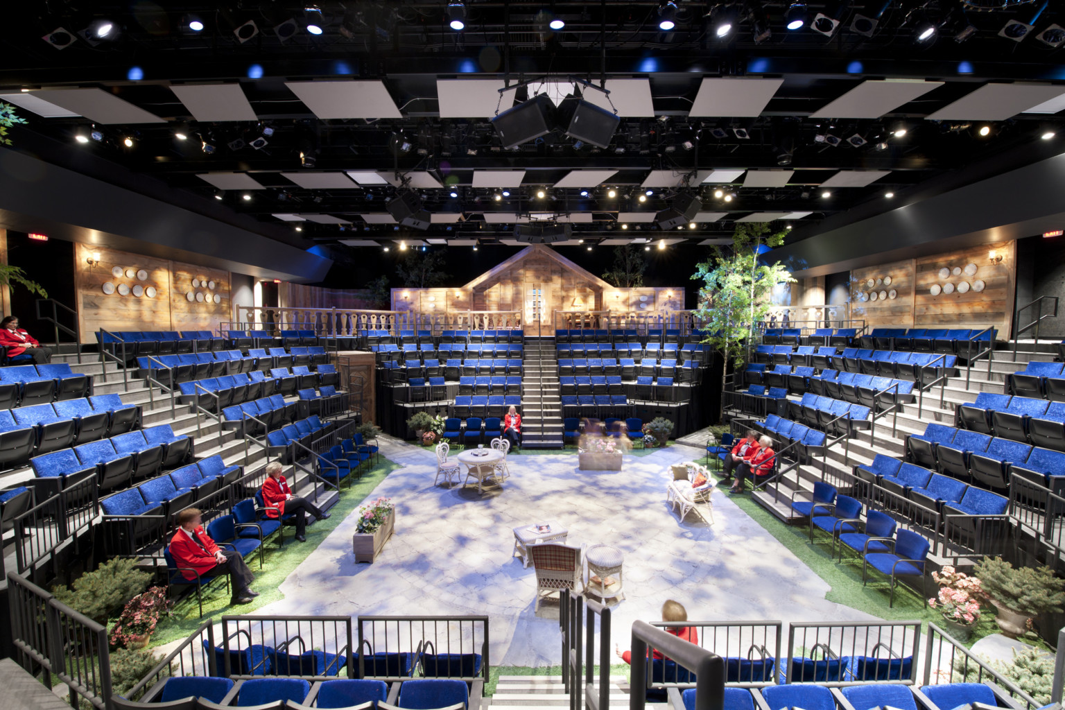 blackbox theater in the round with 360 degree seating around a stage set with tables and chairs, plants at edges