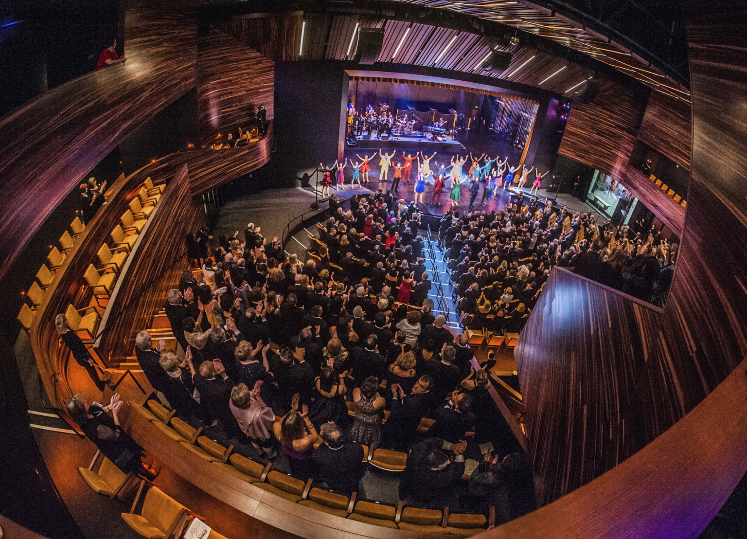 fisheye view of theater with audience and cast bowing on stage, surrounded by dark wood paneling and deep purple lights