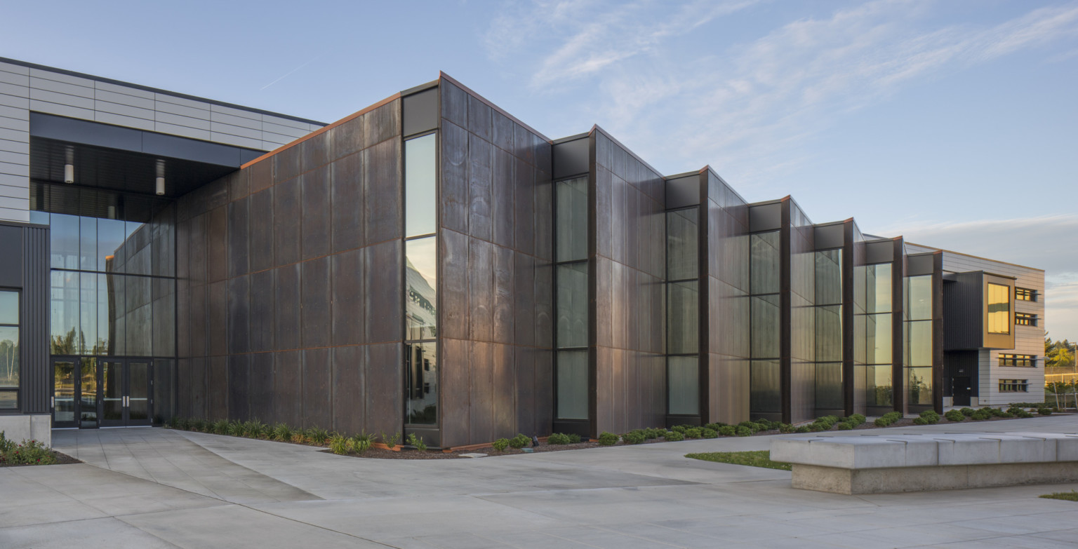 Front entrance of building with brown serrated edge building extensions with strips on windows at end. Double height entrance