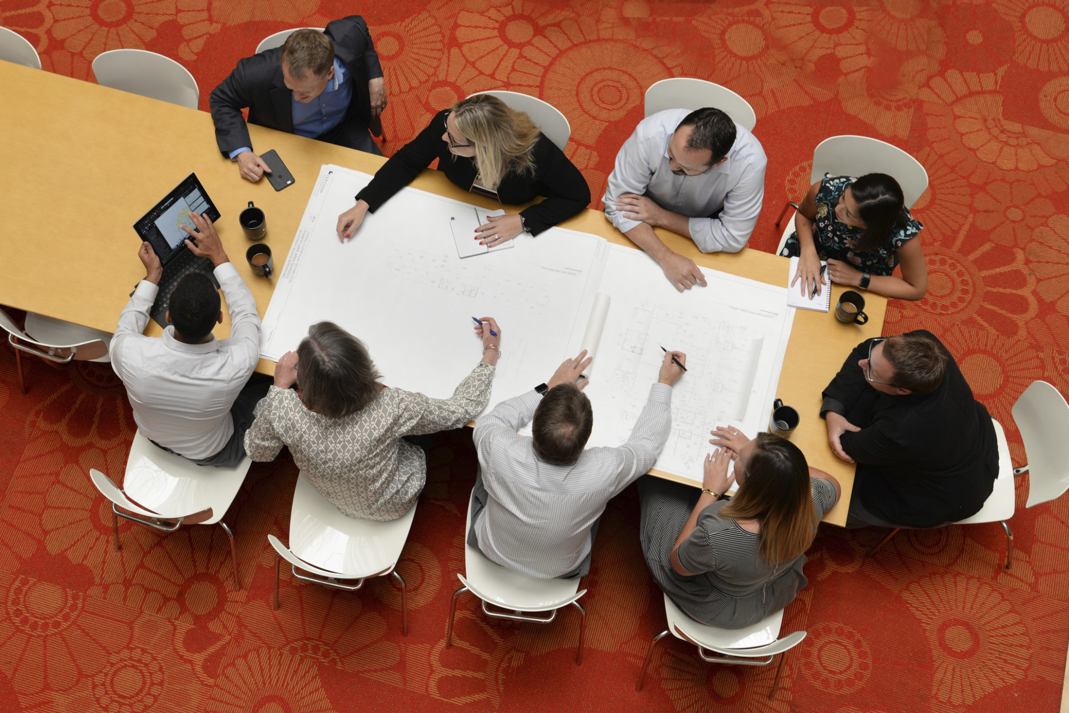 Aerial view of group working at wood conference table with grey chairs on red floral rug. White paper and tablet in use