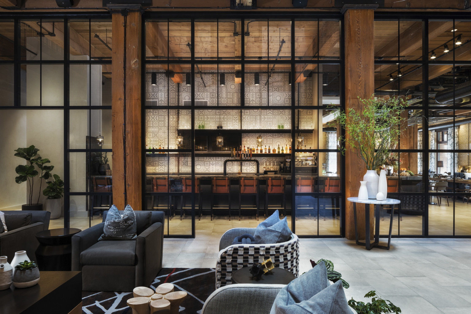 Seating area in room with double height windows looking to white geometric walled bar in next room. Wood columns and ceiling