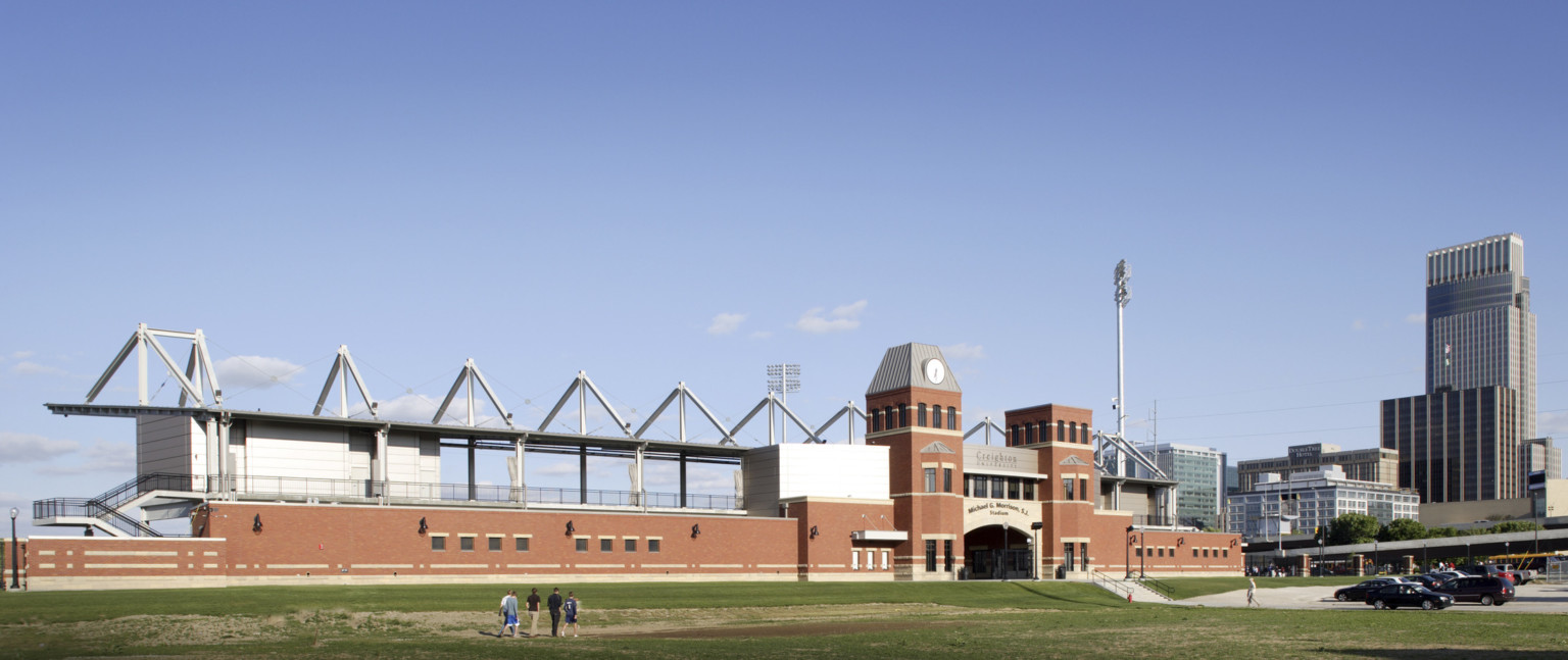 Arched stadium entry framed by brick towers and single story building. Behind, exposed 2nd floor walkway with vaulted beams