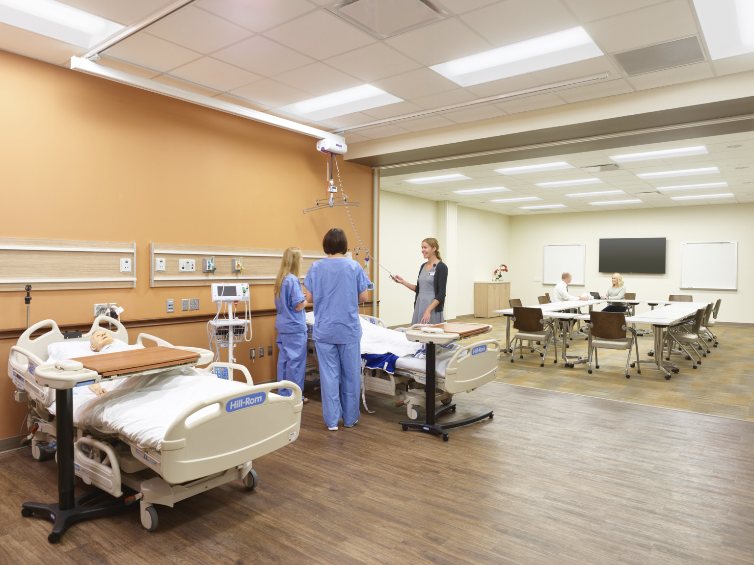 CPR dummies in hospitals beds against brown wall on hardwood floor in front of room. Carpeted back room with flexible seating