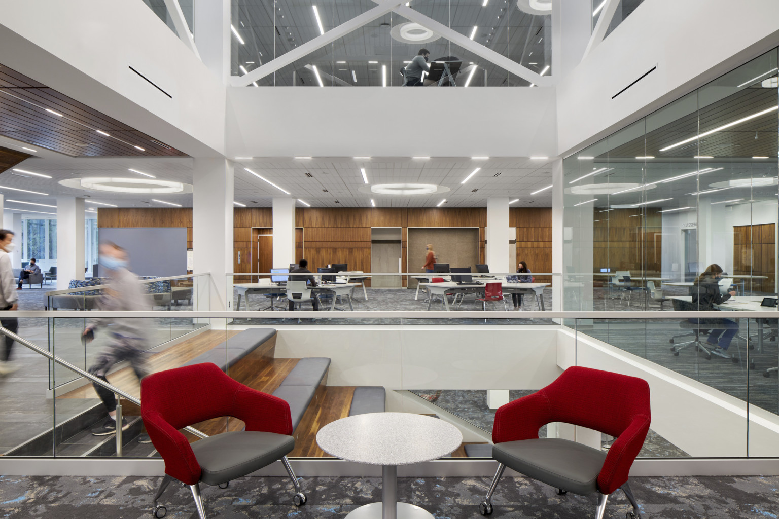 2 red seats in large atrium area with glass balcony separating from stairs. White room with wood paneling behind with seating