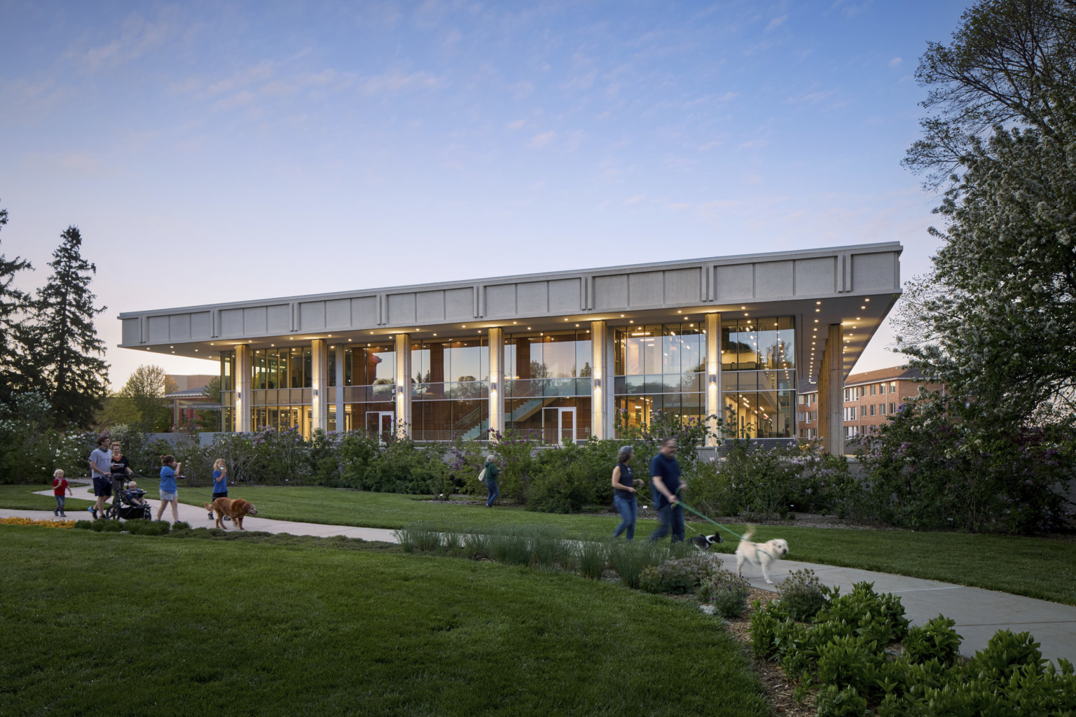 Exterior of a 2 story building with glass windows and cement overhang with columns viewed from across the garden with walkway