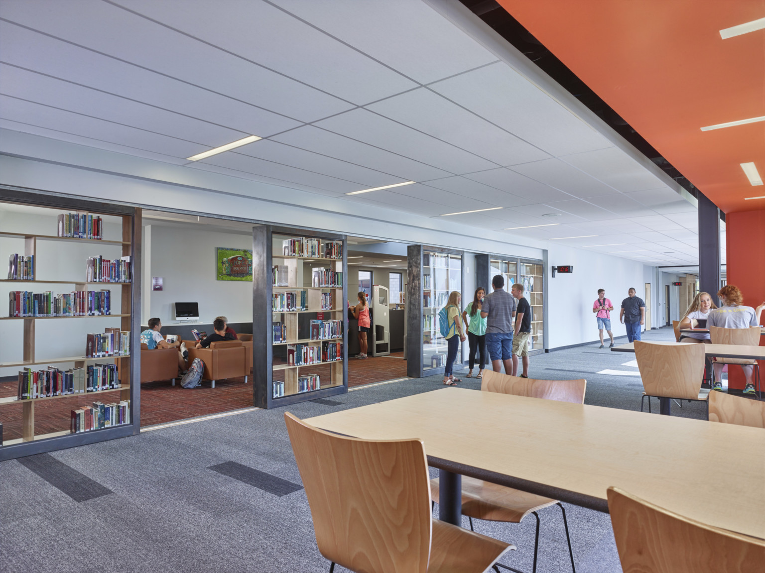 Seating to right framed by orange ceiling accent in white hallway with geometric bookshelves in left wall. More seats beyond