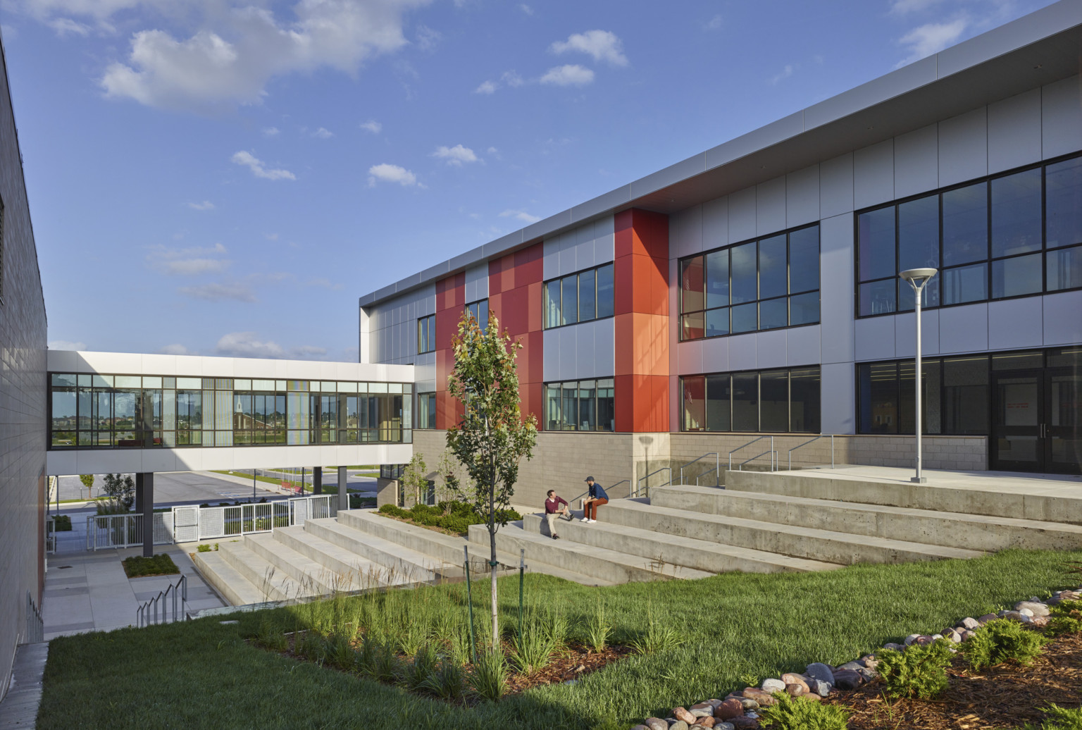 Courtyard with grassy lawn and concrete stairs between 2 white buildings connected by skywalk. Orange accent panels on right