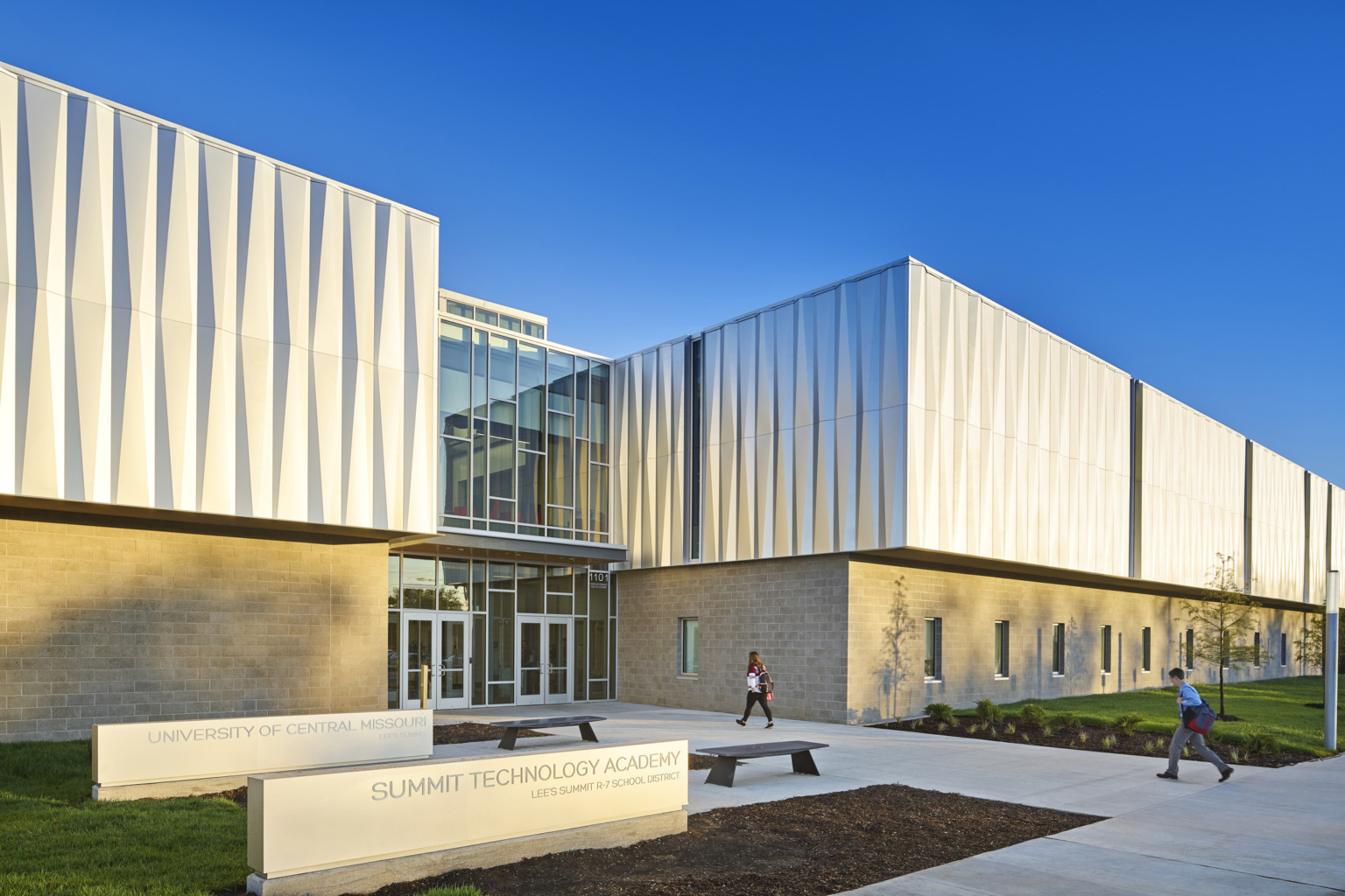 Exterior view of 2 story building with stone lower building and textural metallic upper. Double height windows at center