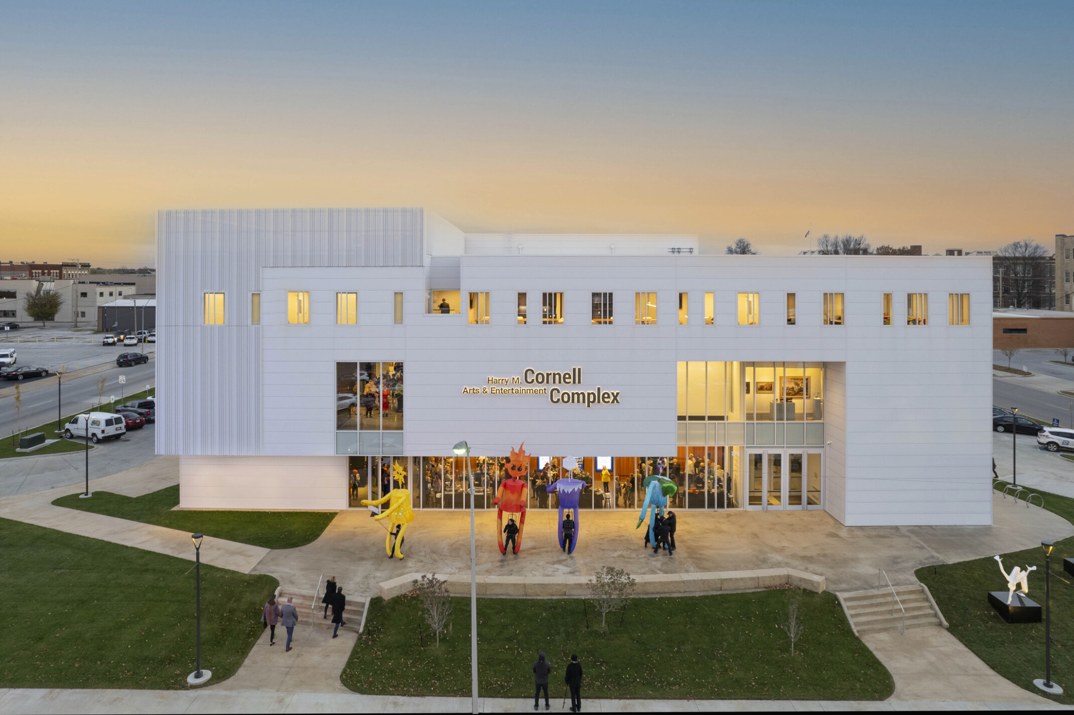 White multistory building light up at dusk by yellow lights; sculptures fill the lawn with people filing in to watch a show at the theater