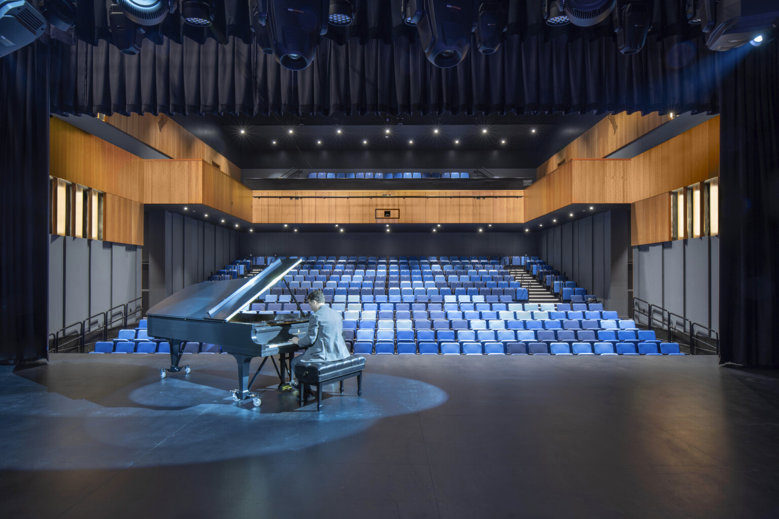 Auditorium theater with piano on stage and pianist performing. Black audience chamber with wood accents and blue seats