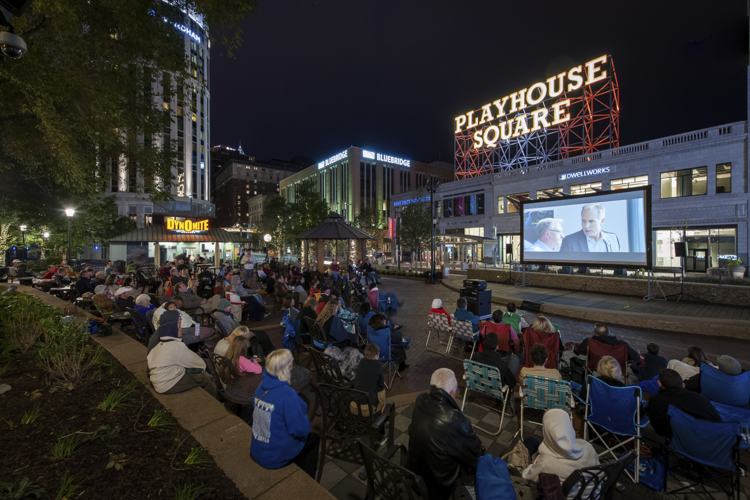 Audience sits on the street watching an outdoor movie on a screen. A Playhouse Square sign is illuminated in the background