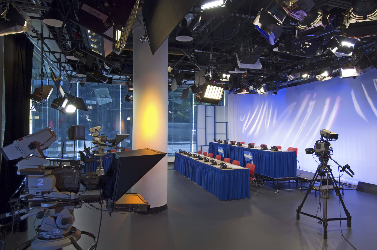 Television studio at street level with three cameras pointed at two rows of tables with chairs and phones, lit from above