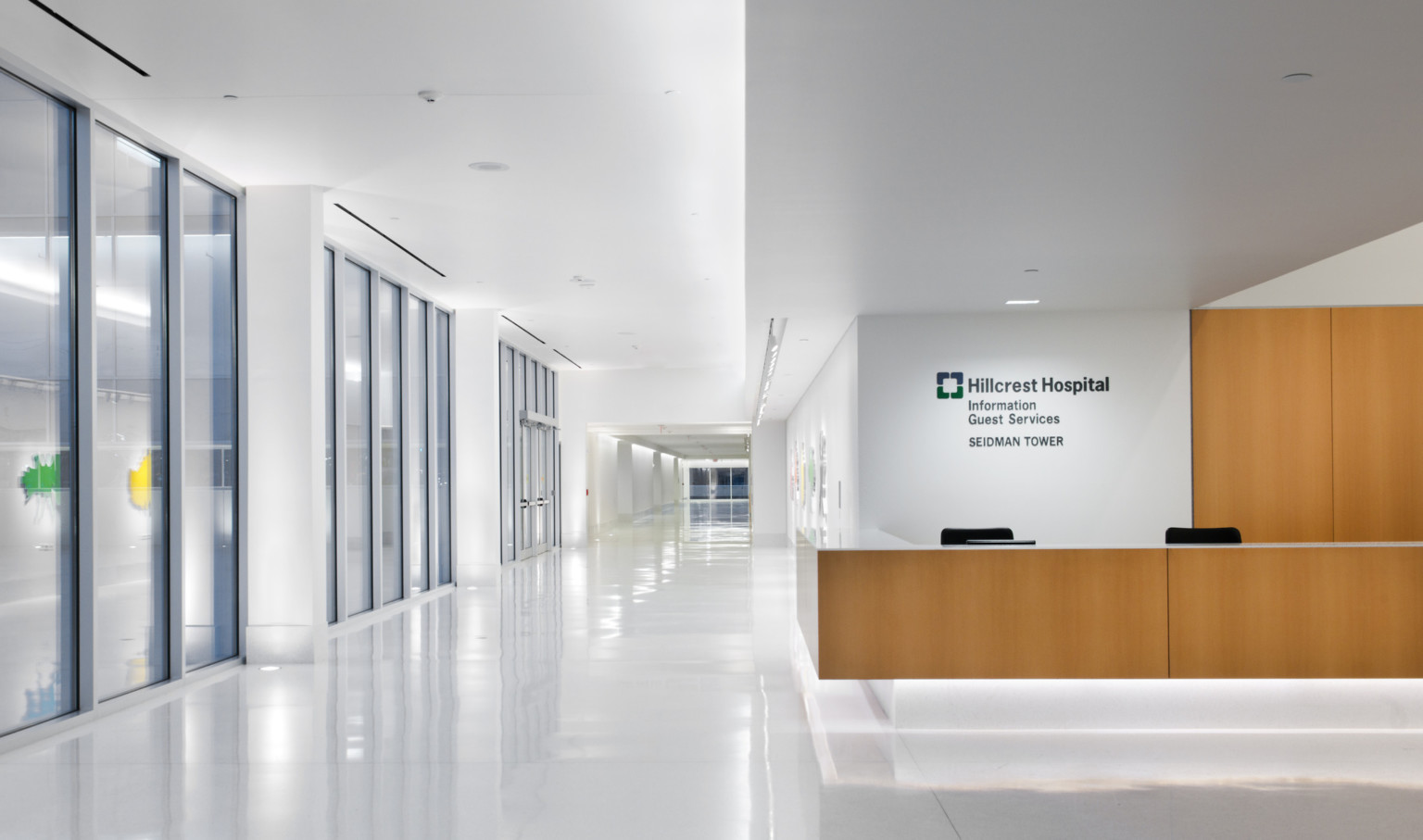 White hallway with wood accent reception desk, wall above reads Hillcrest Hospital Information Guest Services Seidman Tower