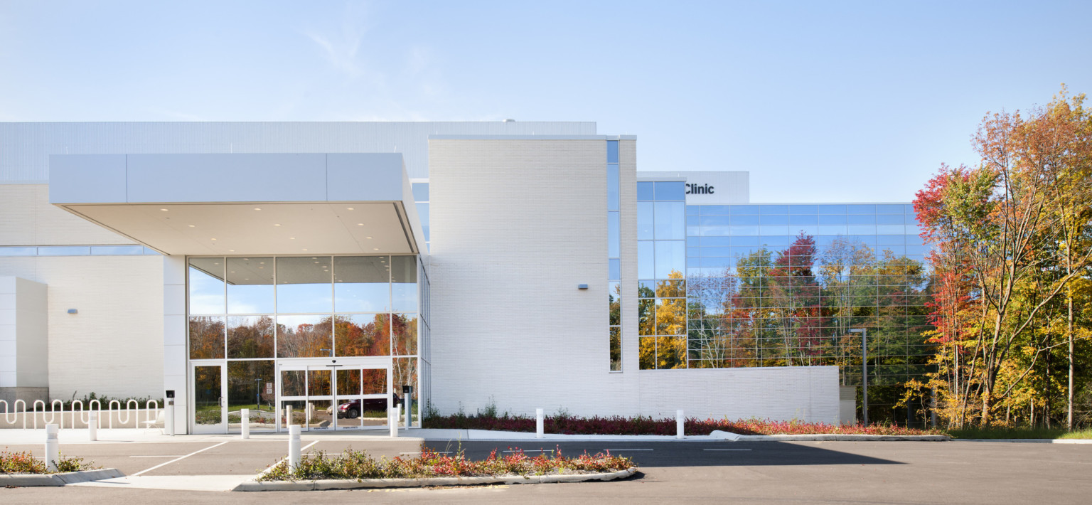 Entry with double height windows with white overhang extending over driveway. White wall right with window wall beyond