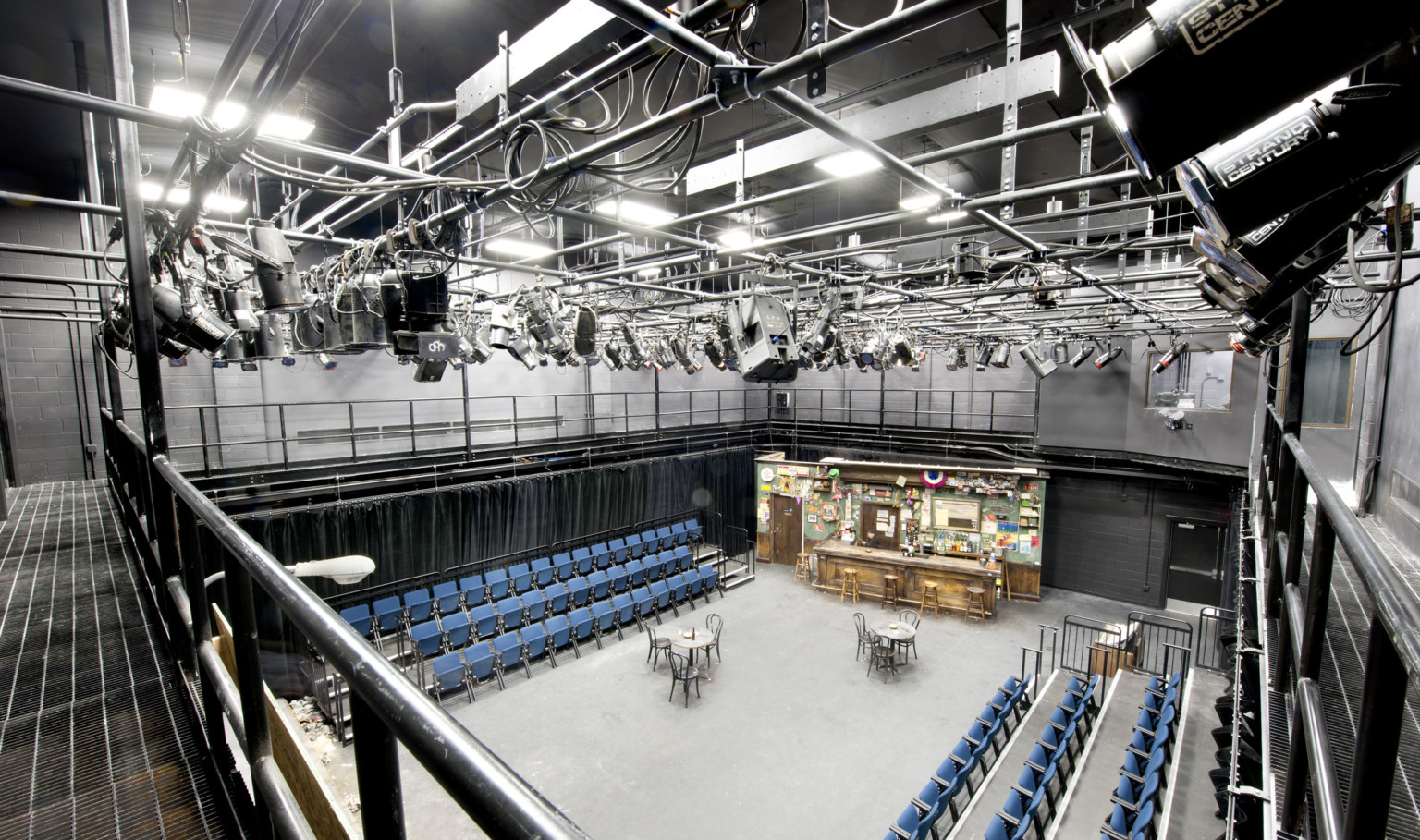 Black box theater with side seating, lights hang on a metal grid above. The stage is set with tables, chairs, and a bar