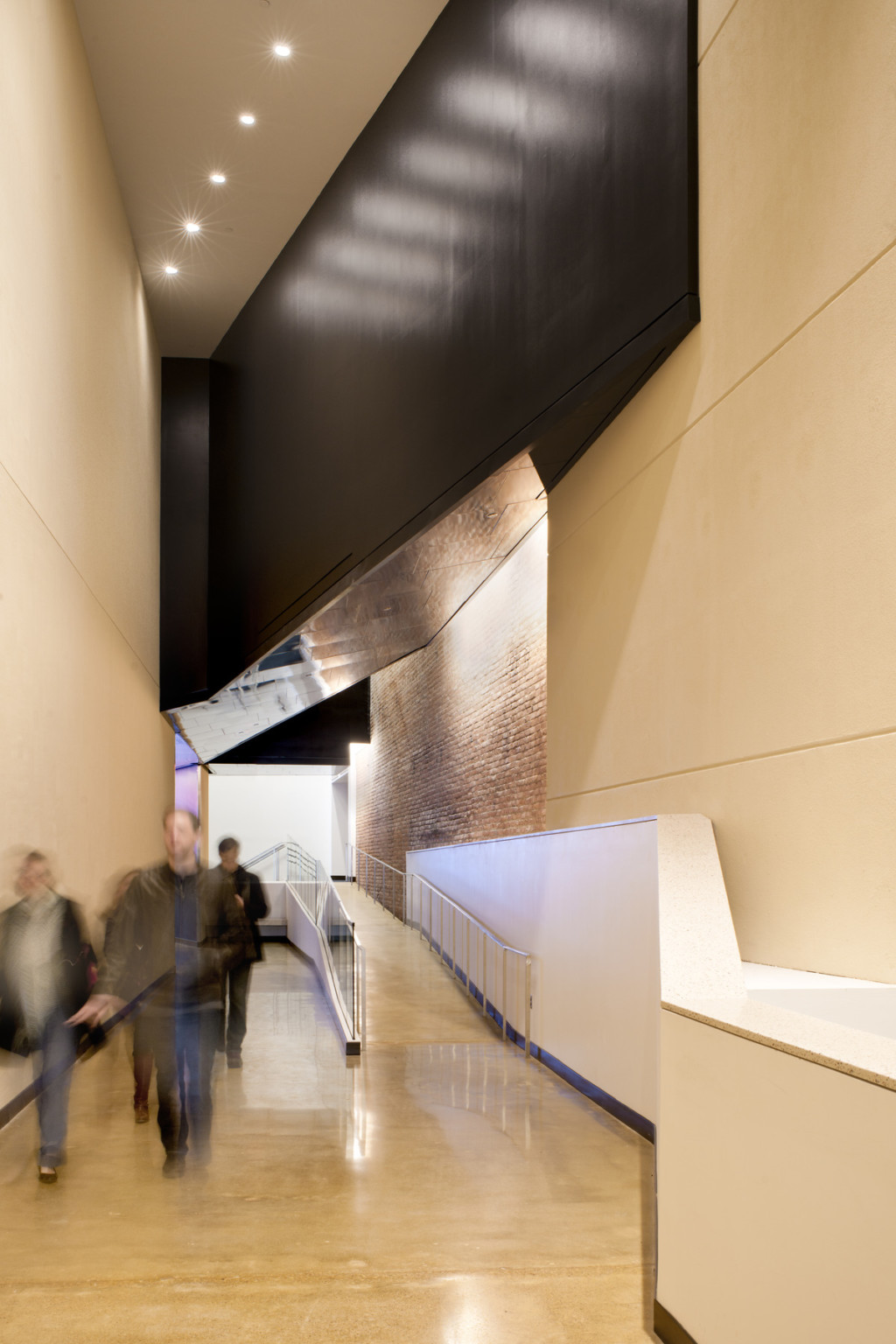 Hallway with ramp to the right, concrete yellow walls with exposed brick section reflected on metal ceiling section