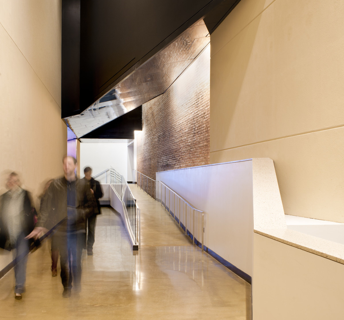 Hallway with ramp to the right, concrete yellow walls with exposed brick section reflected on metal ceiling section