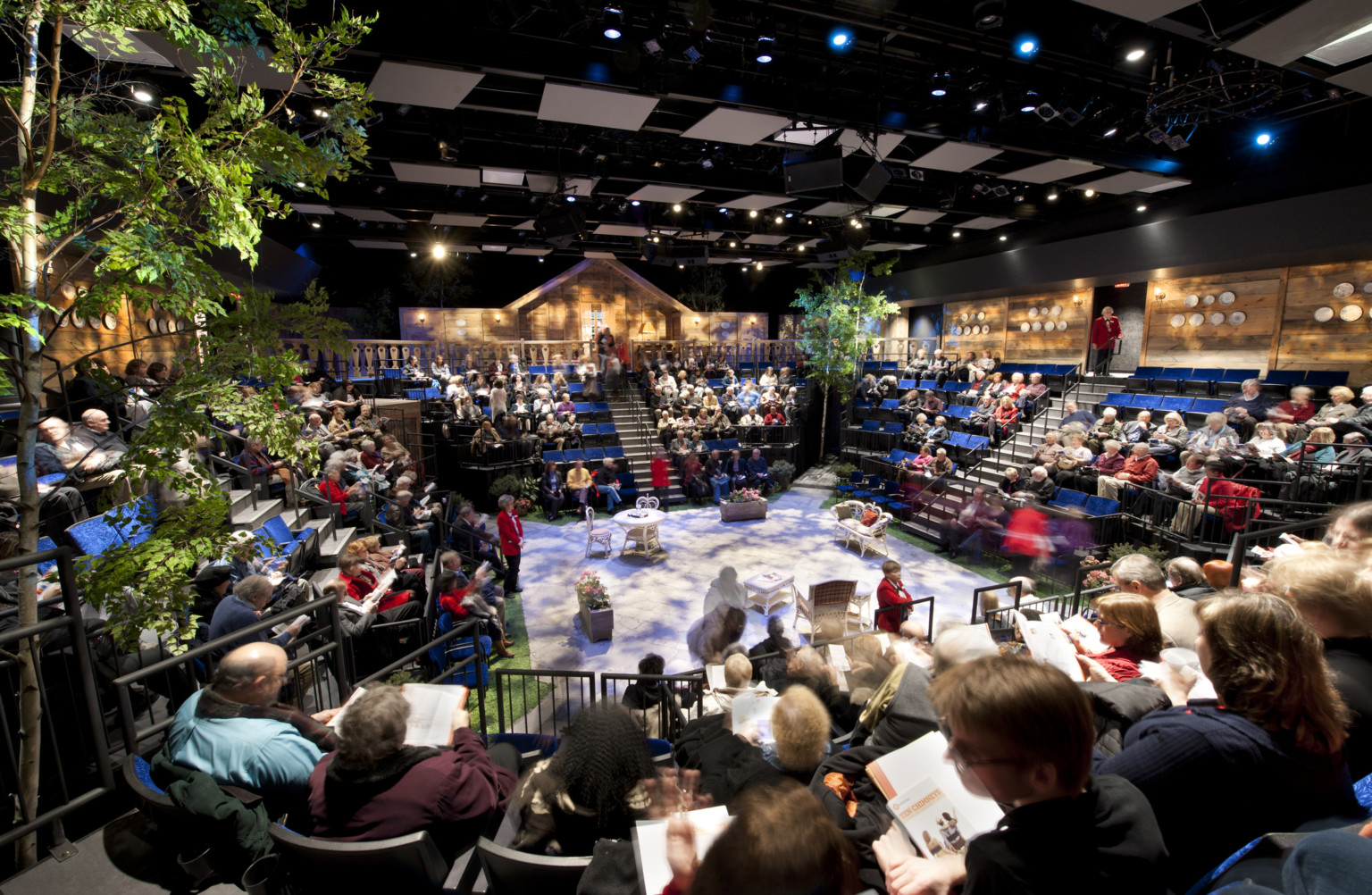 people seated at theater in the round with trees off a stage set with white table and chairs, textural lighting from above