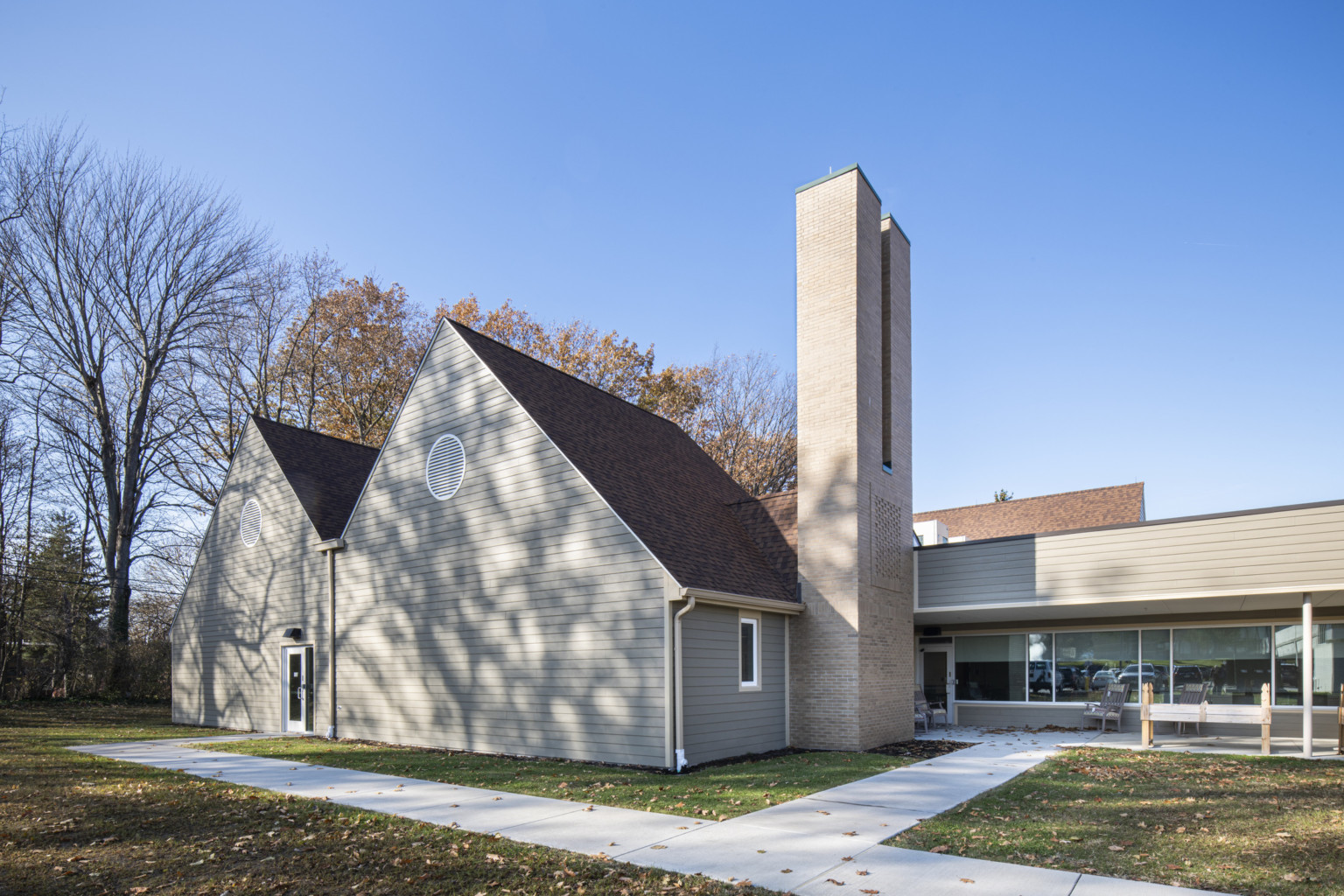 Gable framed building with tan clapboard siding and tan brick chimney. concrete path walkways lead to glass front entry