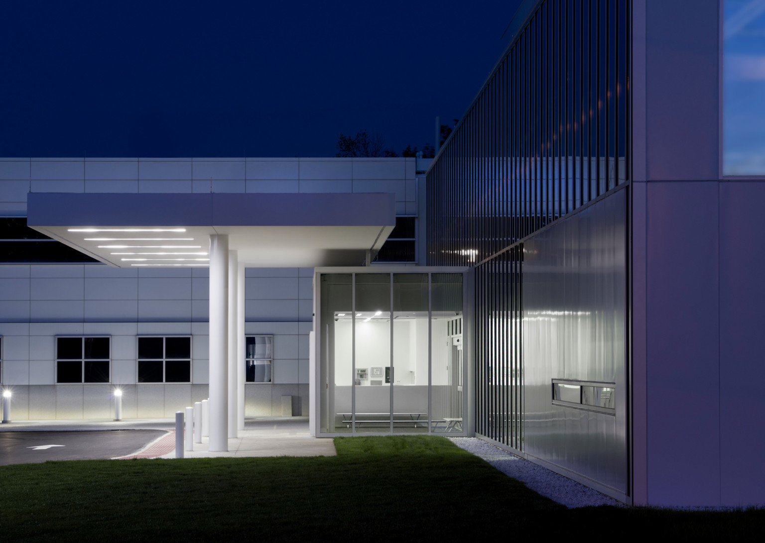 Side view of entry with glass vestibule with benches. Overhang above entry to driveway. Grey wall behind with square windows