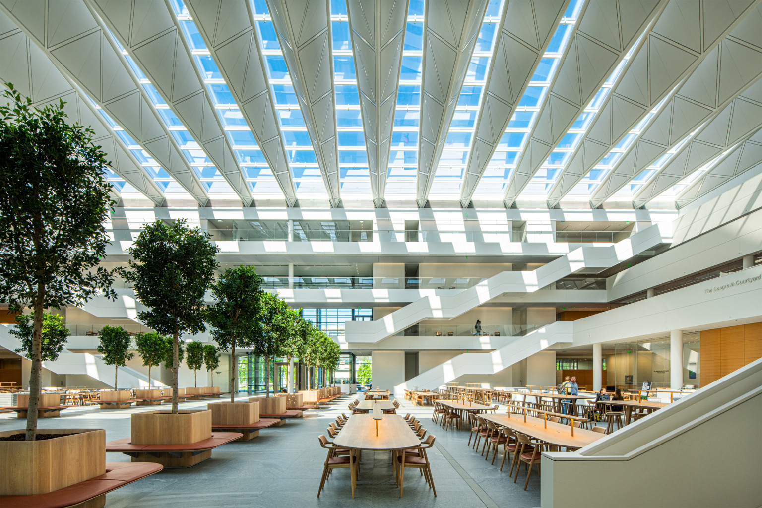 Atrium seen from ground level. Long tables with chairs on either side of tree lined walkway. White angular staircases at back