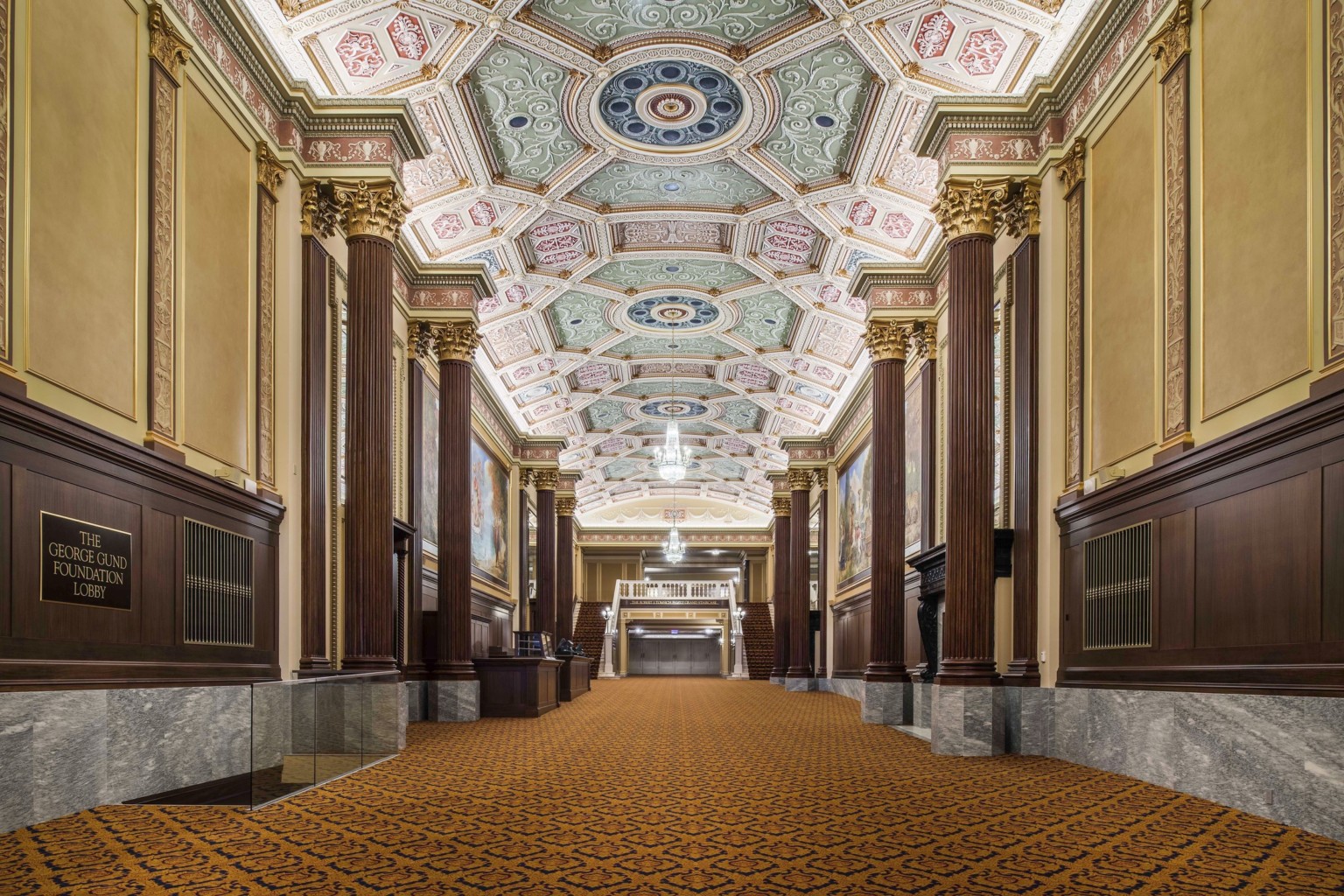 Grand staircase carpeted beneath ornate multicolored coffered ceiling with chandelier framed by ribbed pillars