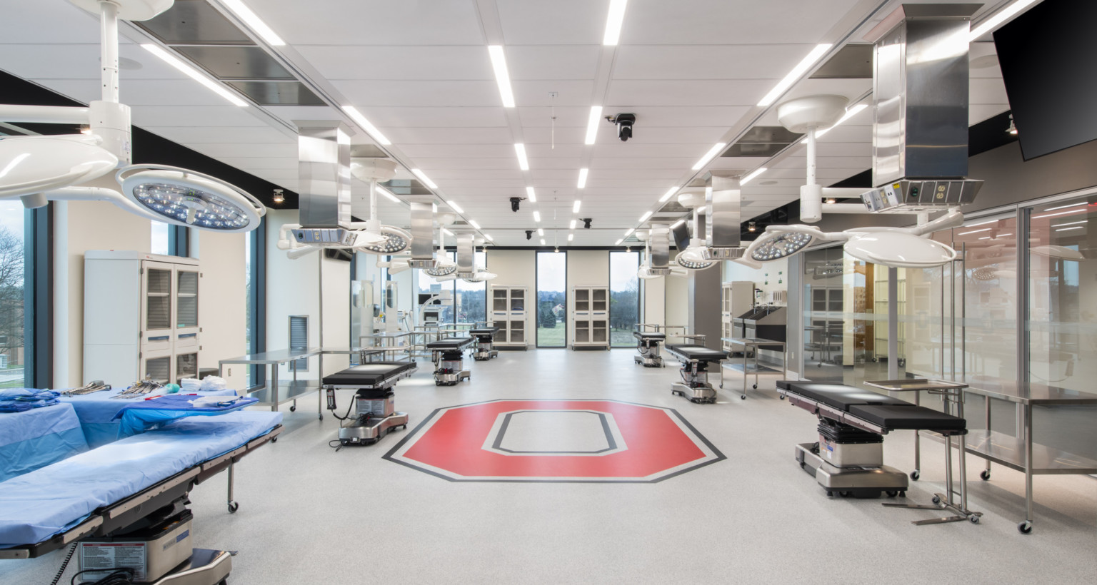 Inside view of room with 6 hospital beds with 2 circular lights hung above, tables next to the bed and red O on floor