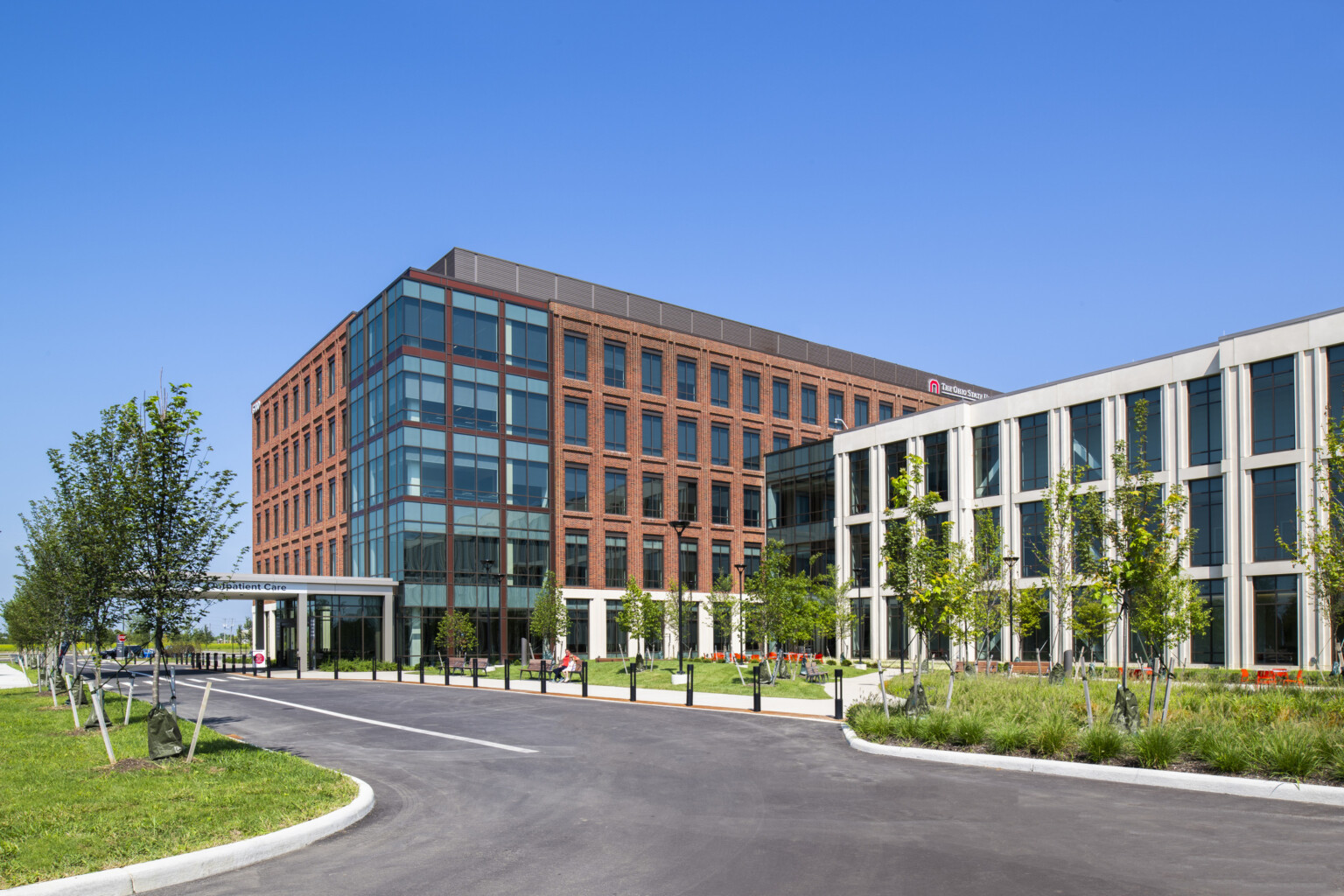 Brick building with glass corner extending past connected shorter long white building with large windows behind courtyard