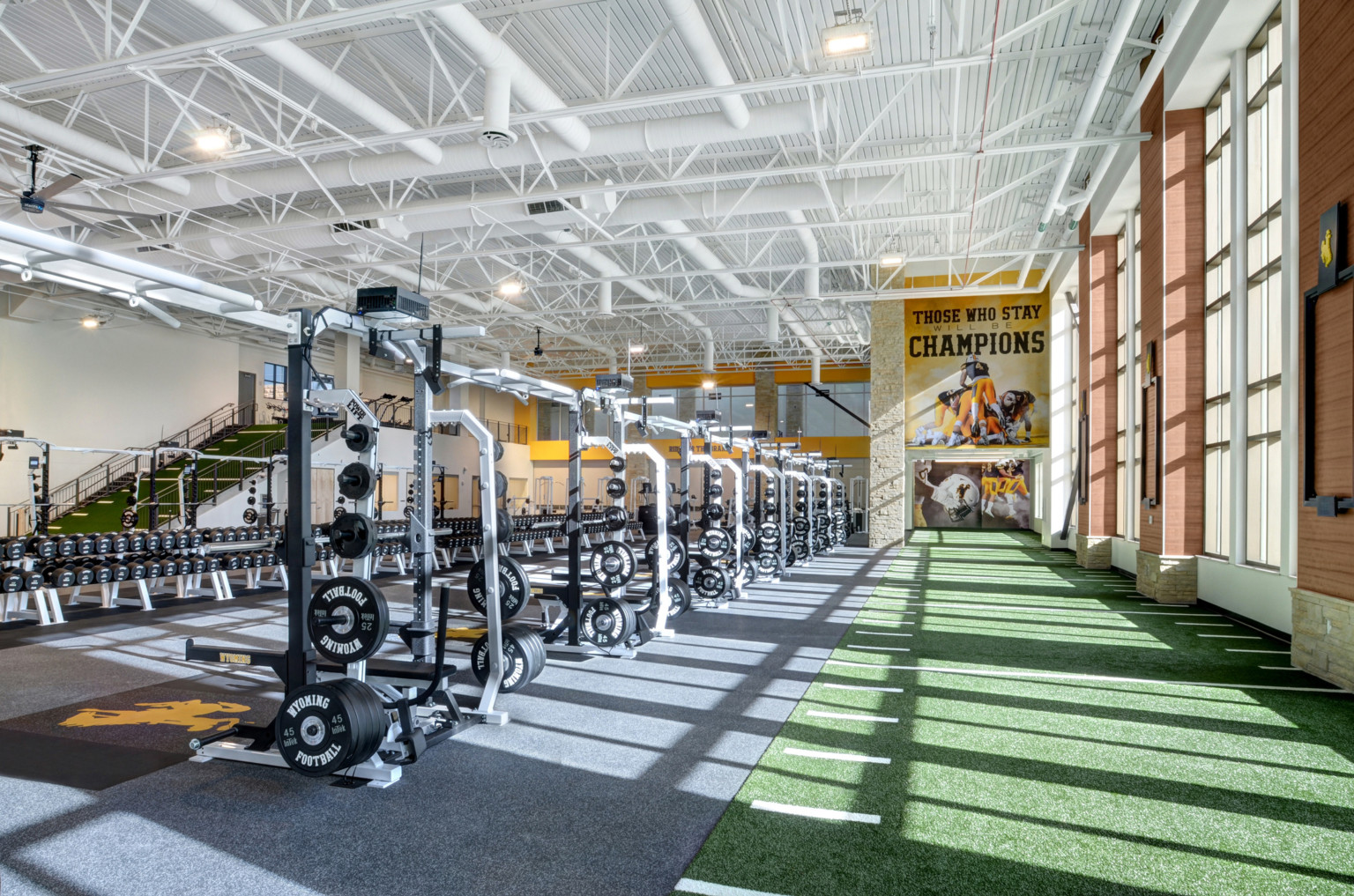 Turf floor with yard marks along double height windows. Ahead, a mural reading Those Who Stay Will Be Champions. Weights left
