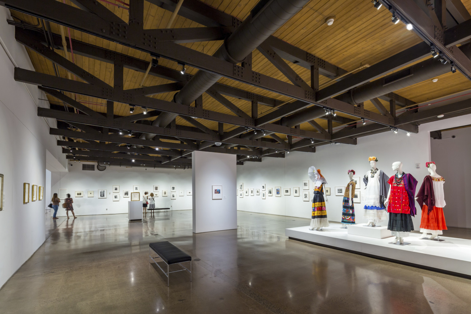 A gallery with paintings and a display of dressed mannequins, under a wood ceiling with exposed black support beams and pipes